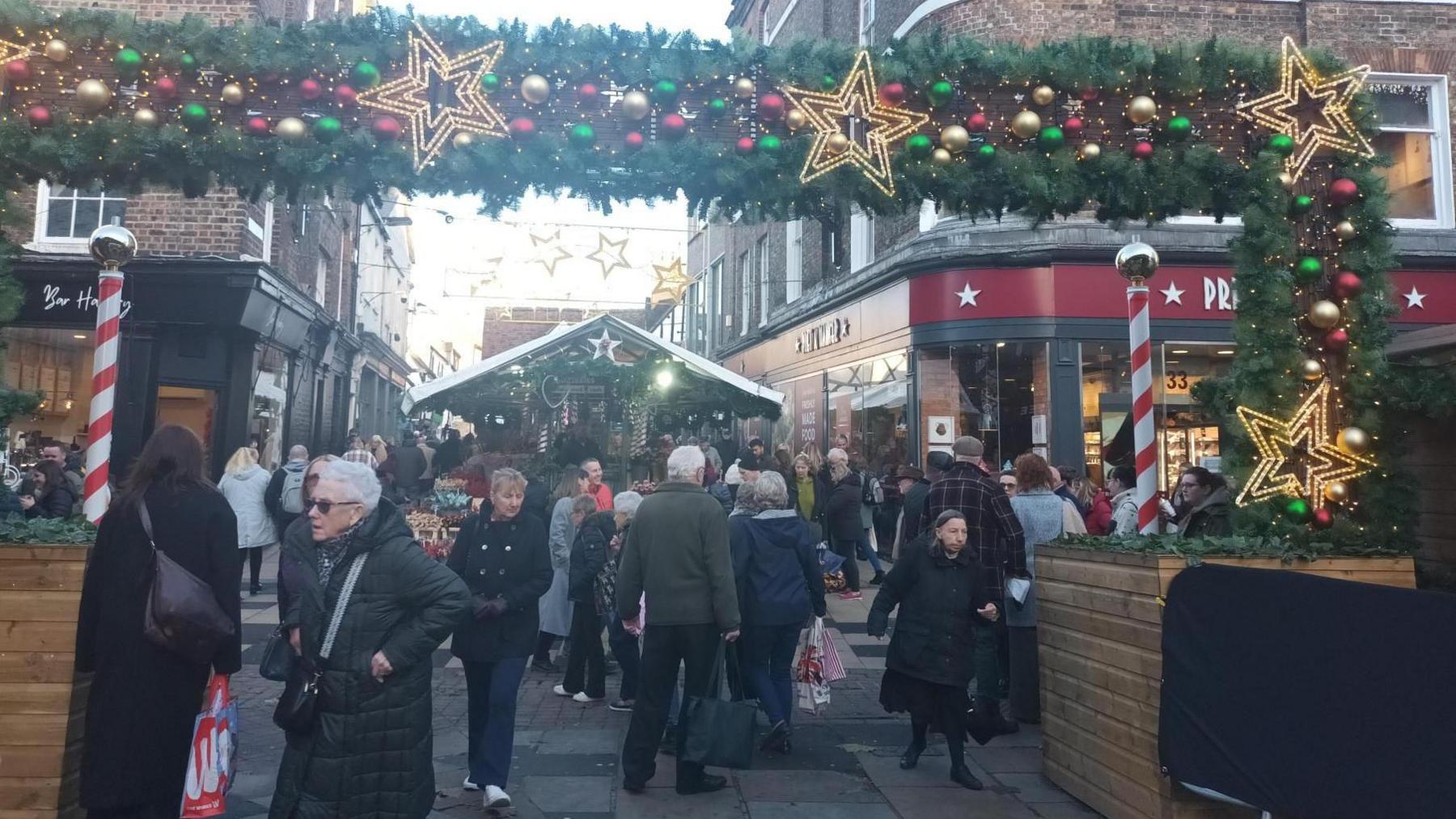 A shot of the Christmas Market on Parliament Square in York, with a large fir tree archway, decorated with gold, green and red baubles and illuminated stars. A mixed crowd of people can be seen walking beneath it.