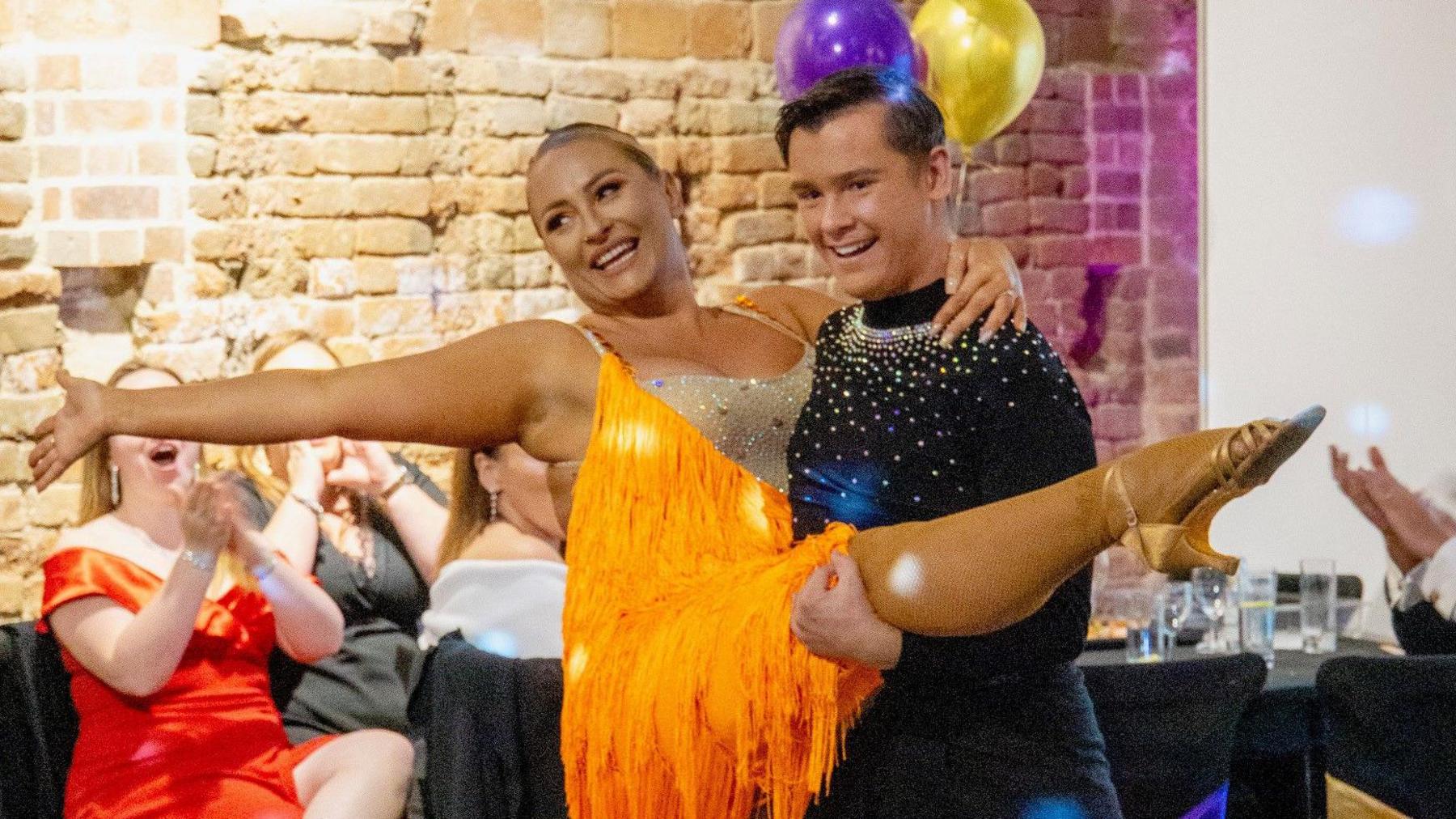 A man wearing a black outfit with sparkles on it lifting a woman who wears an orange filly dress