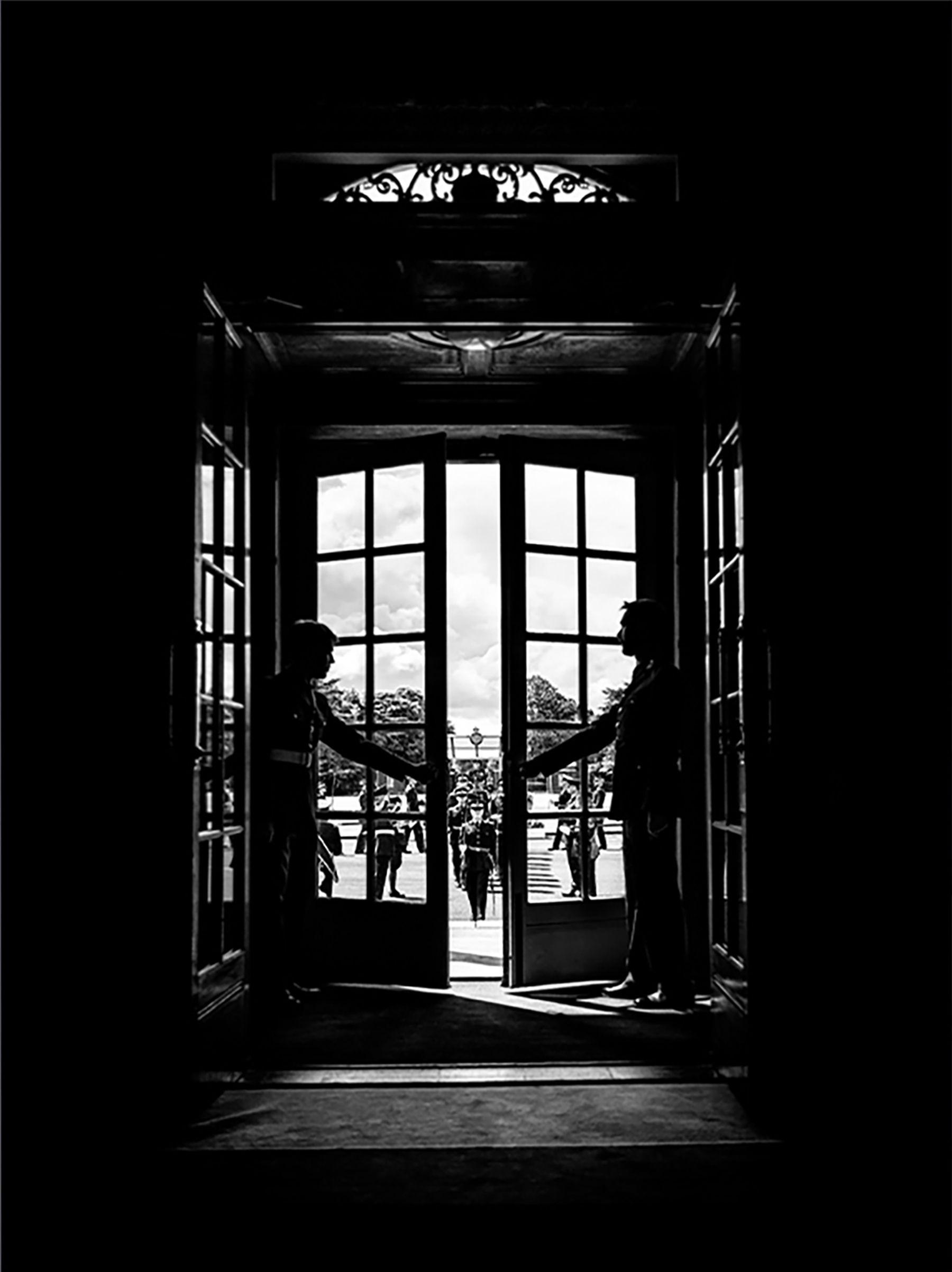 Two officers hold the doors in waiting to open them to the trainees on their graduation day.  The black and white photo is mostly black and the officers are in the distance through the window.