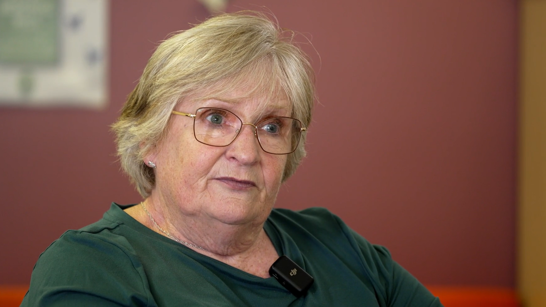 An older woman with short blonde hair is seated indoors, wearing a green top with a small microphone clipped to the collar. She is wearing glasses. The background is softly blurred, featuring a purple wall and indistinct objects.