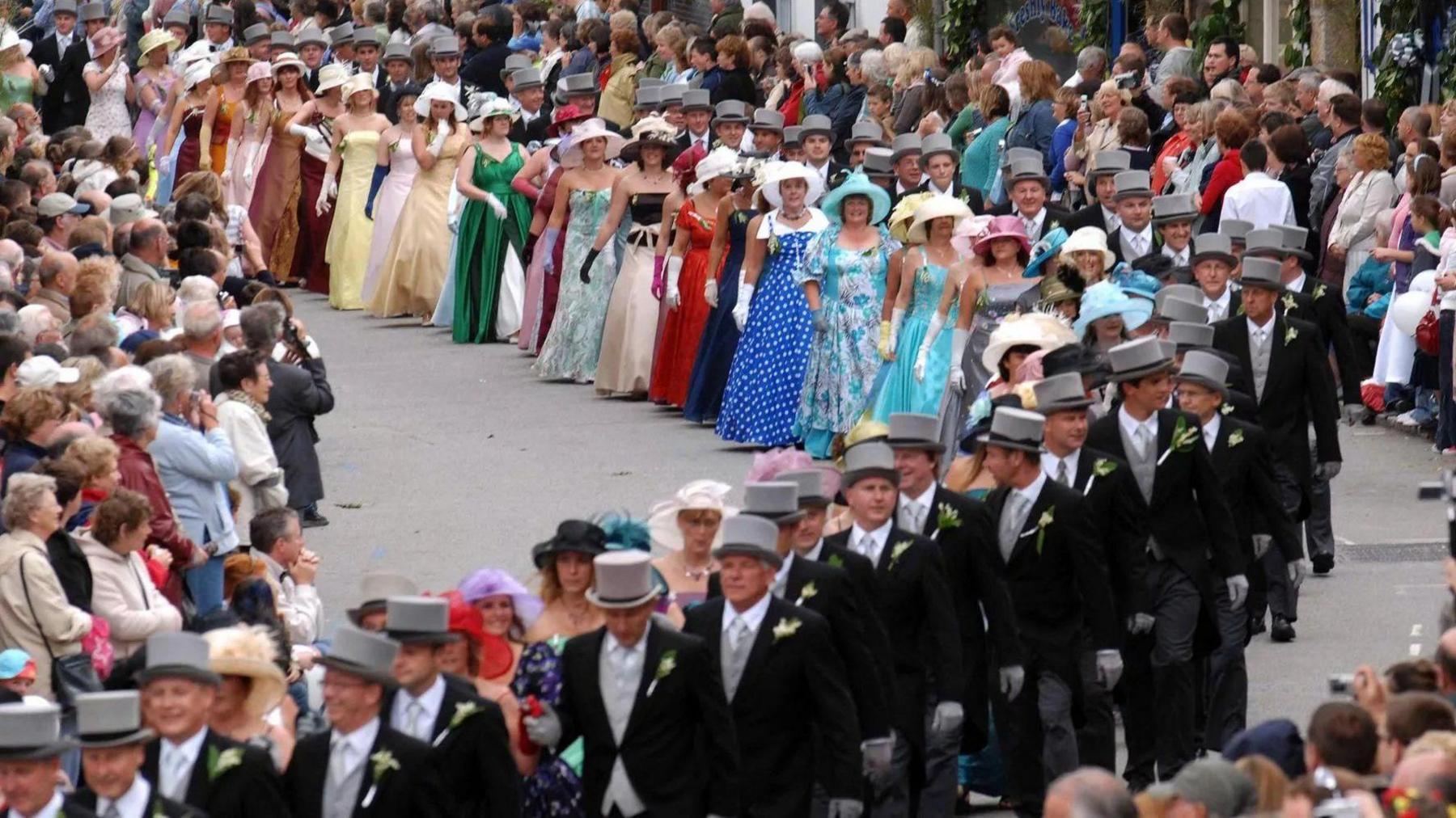 About 30 couples are walking along a street which is lined with spectators. The ladies are wearing brightly-coloured full-length gowns with hats and gloves. The gentlemen are wearing top hats and tails. The shops lining the street are decorated with greenery and flowers.