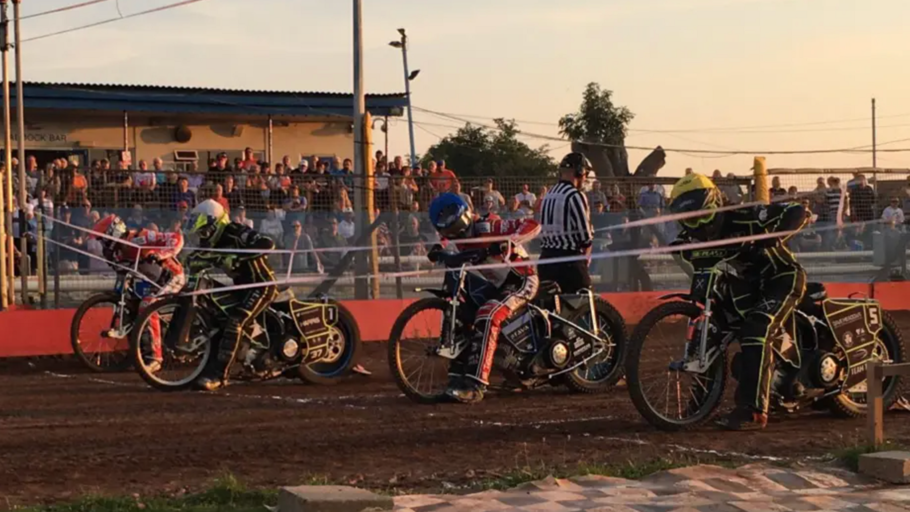 There are four motorcyclists on the starting line of the dirt track at Swindon speedway. A man wearing a black and white striped top is in the centre of the bikes and spectators can be seen in the background.