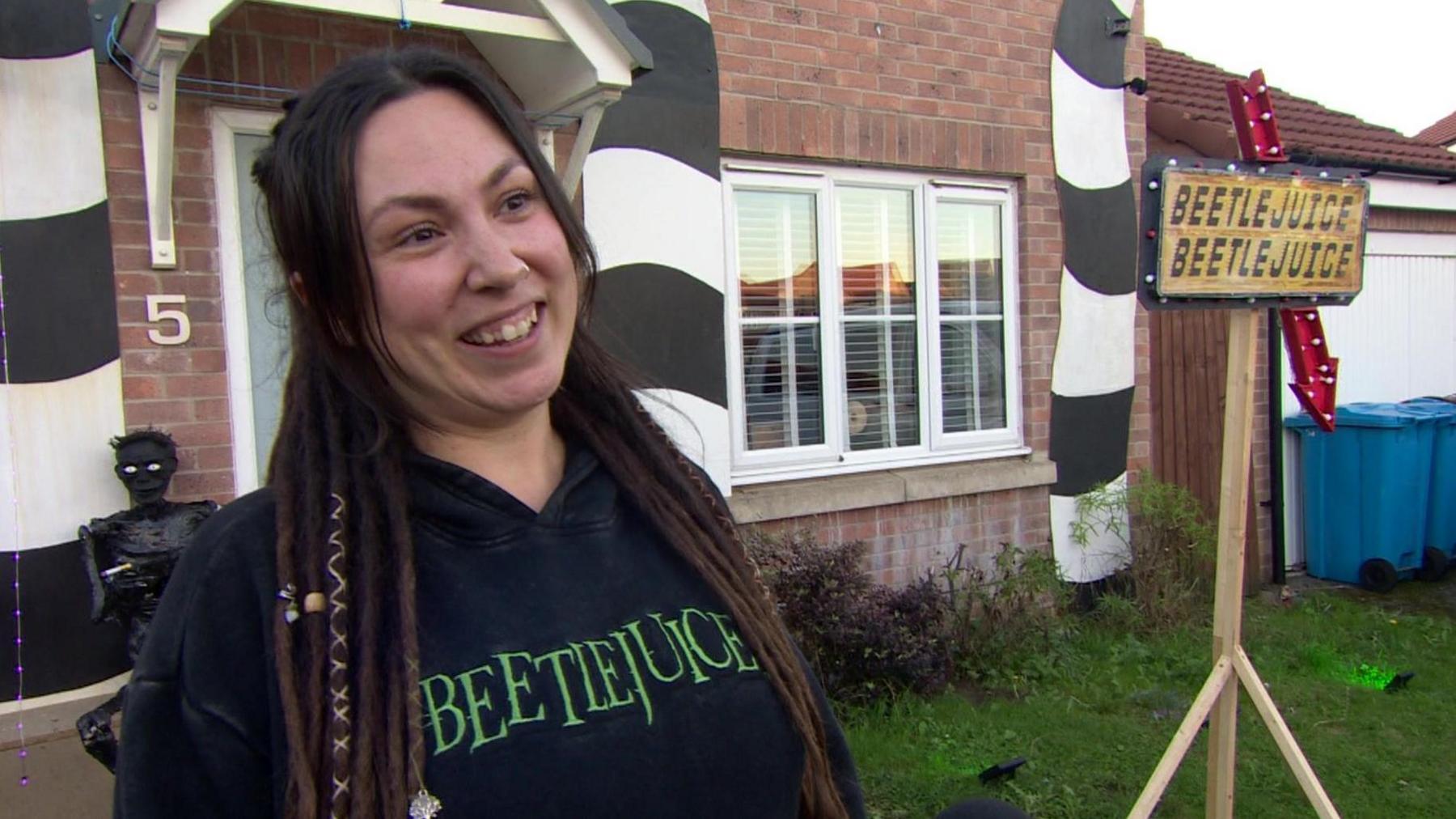 A woman with long dark hair smiles as she is interviewed by the BBC. You can see her Halloween decorations in the background, and she wears a black Beetlejuice hoodie. 