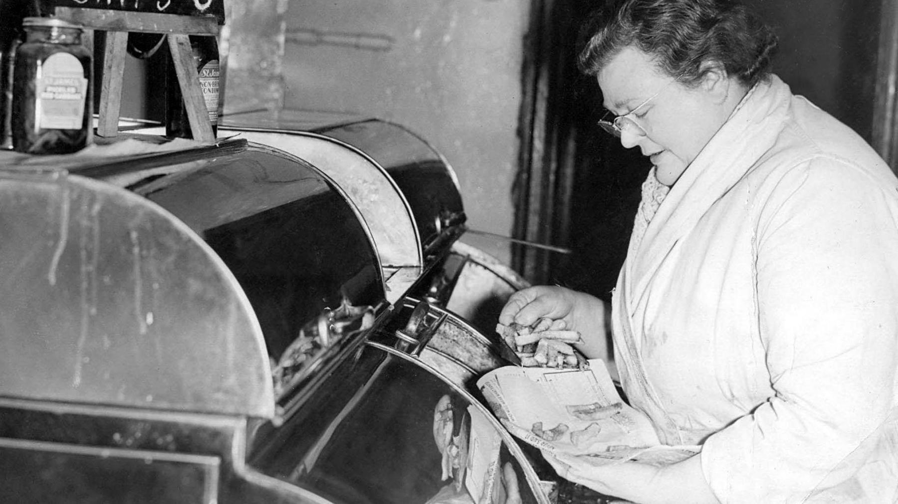 A black and white image, taken in 1956 in Newcastle, showing a woman wrapping chips, fresh out of the fryer, in a sheet of newspaper.