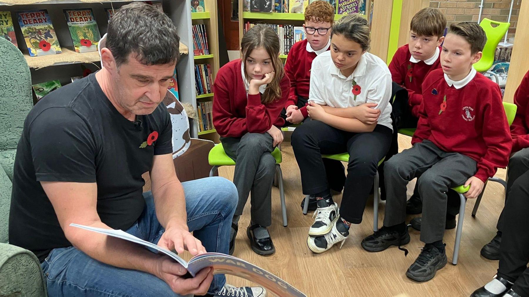 Tom is seated in a library or reading area, holding an open book and reading to a group of children. The children are sitting on green chairs arranged in a semicircle, wearing red school uniforms with white shirts. Some of them have red poppy pins on their clothing.
