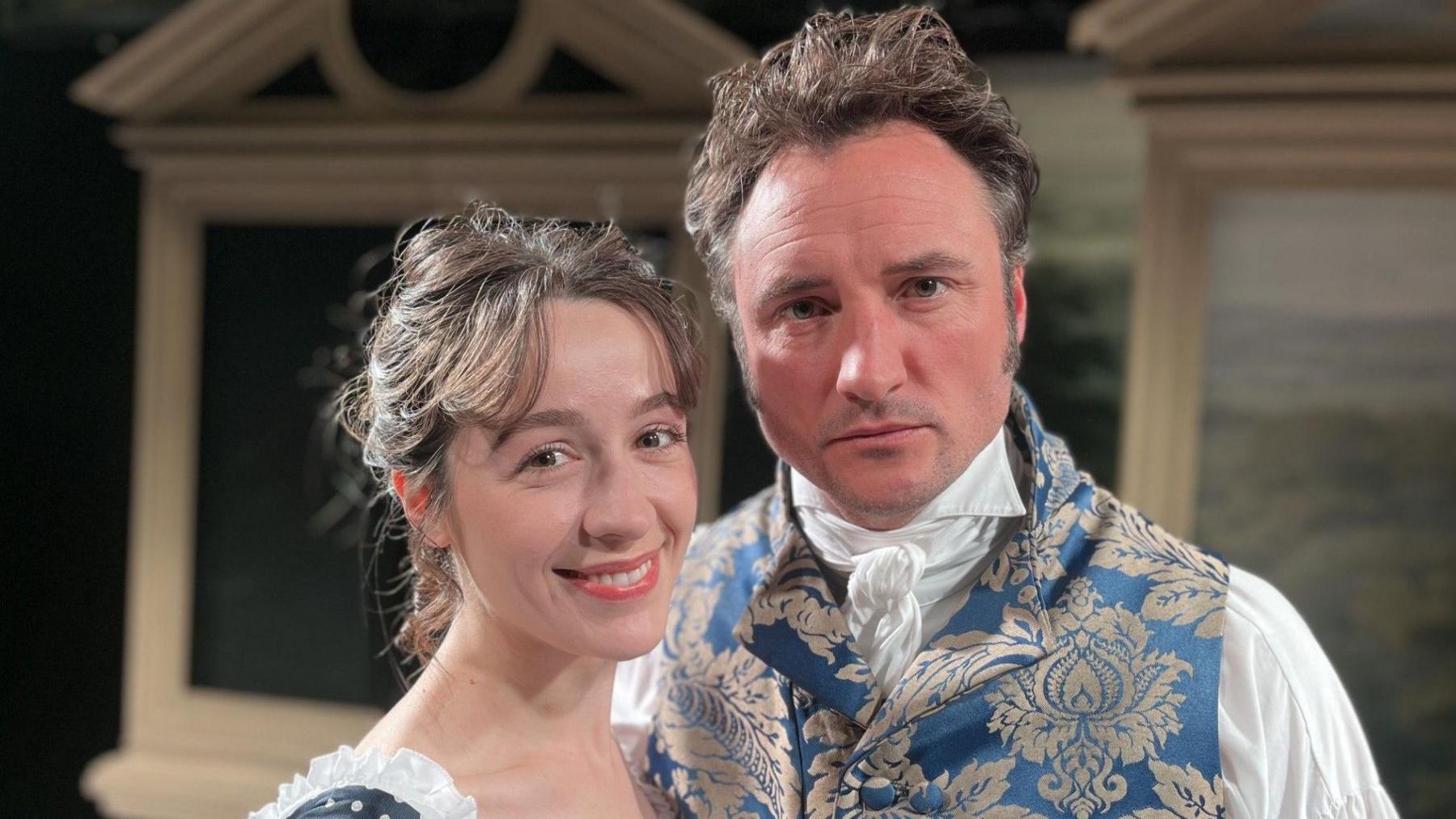 A woman in an old-fashioned dress smiling and a stern-looking man in a patterned waistcoat