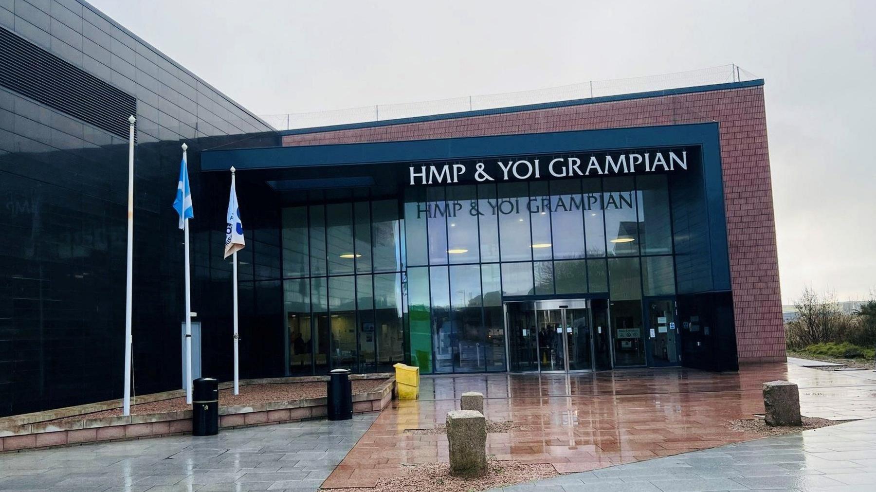 Brick and glass building with sign wording saying 'HMP & YOI Grampian', and flagpoles at the side.