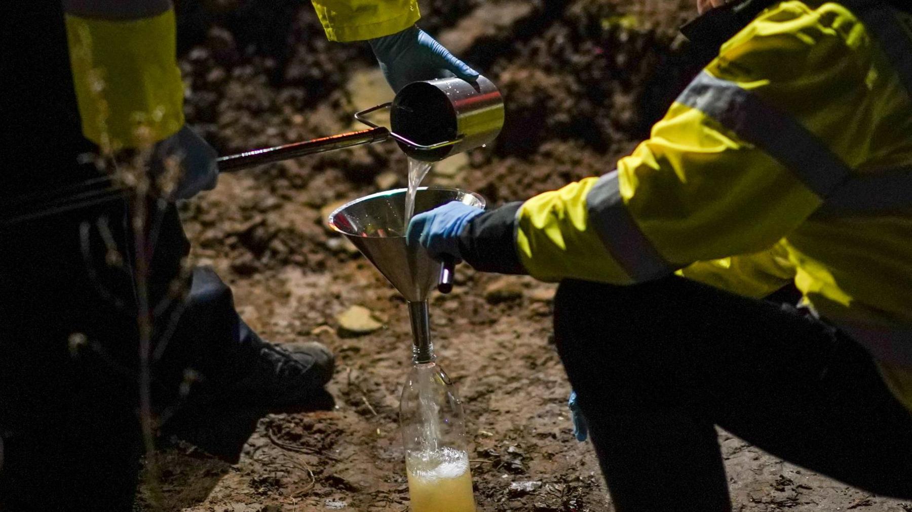 Two workmen in hi-vis jackets sift water into a bottle through a funnel. They appear to be working on a sandy surface.