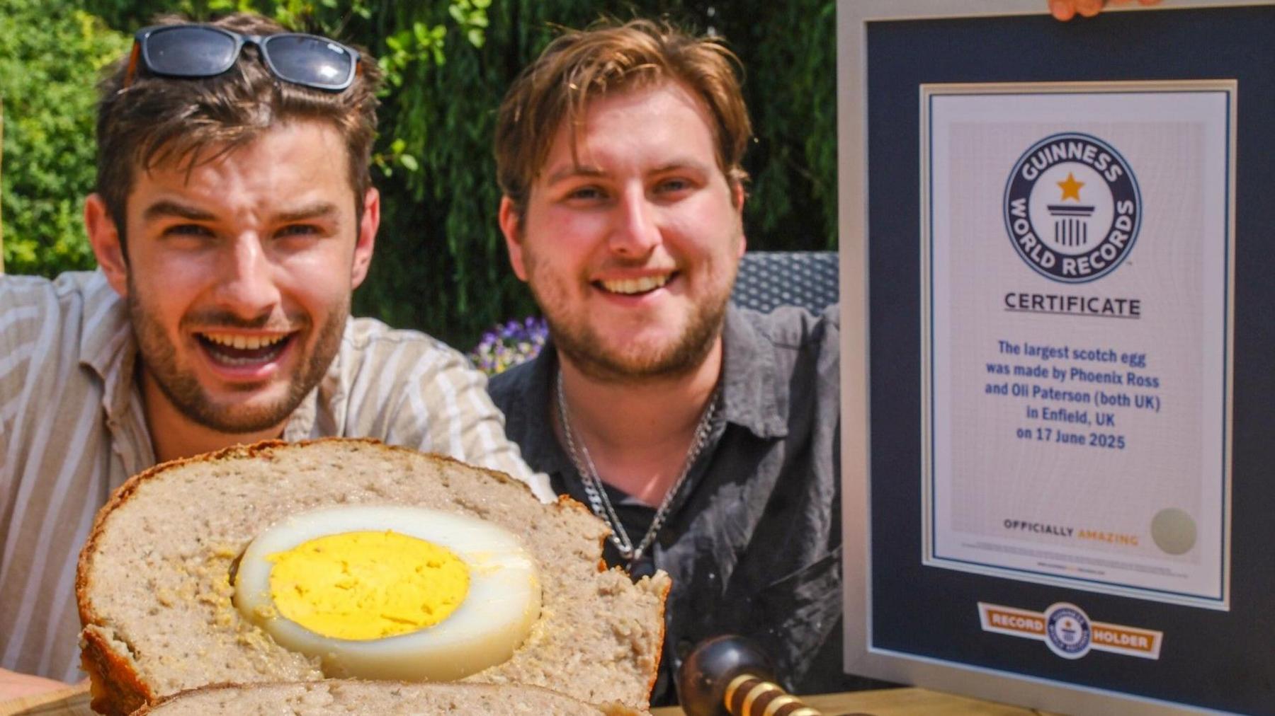 Two men, both with brown hair and short beards, are seated at an outdoor table holding a Guinness World Records certificate and are smiling at the camera. In front of them lies a massive Scotch egg, sliced in half to reveal the egg inside.