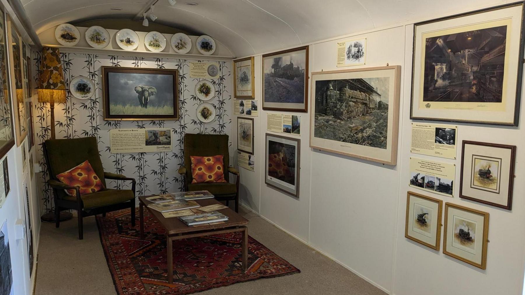 The inside of a repurposed railway carriage showing two chairs at the end with a coffee table and rug, surrounded by paintings and prints on the walls