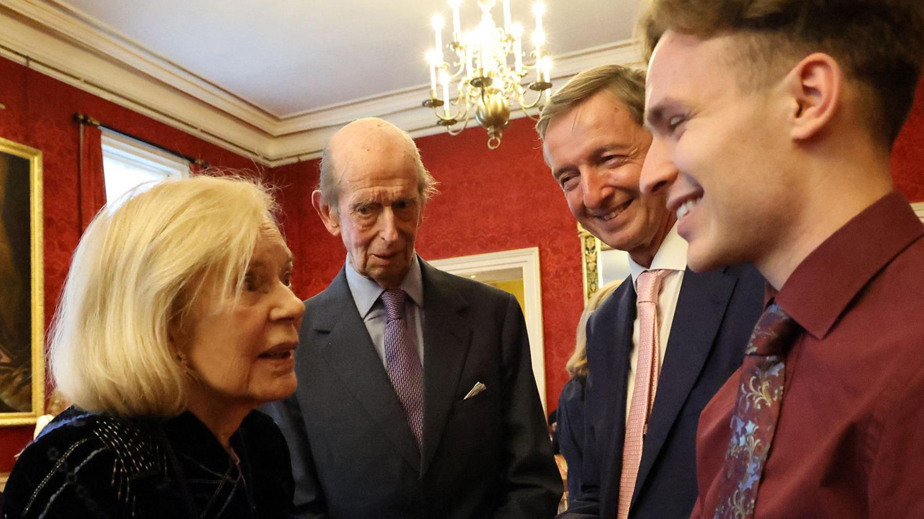 An elderly blond woman looks intently at a young man, most of them are cropped from the shot, but they look like they may be shaking hands. He is smiling widely. Two men stand the other side of them looking on, towards the camera.