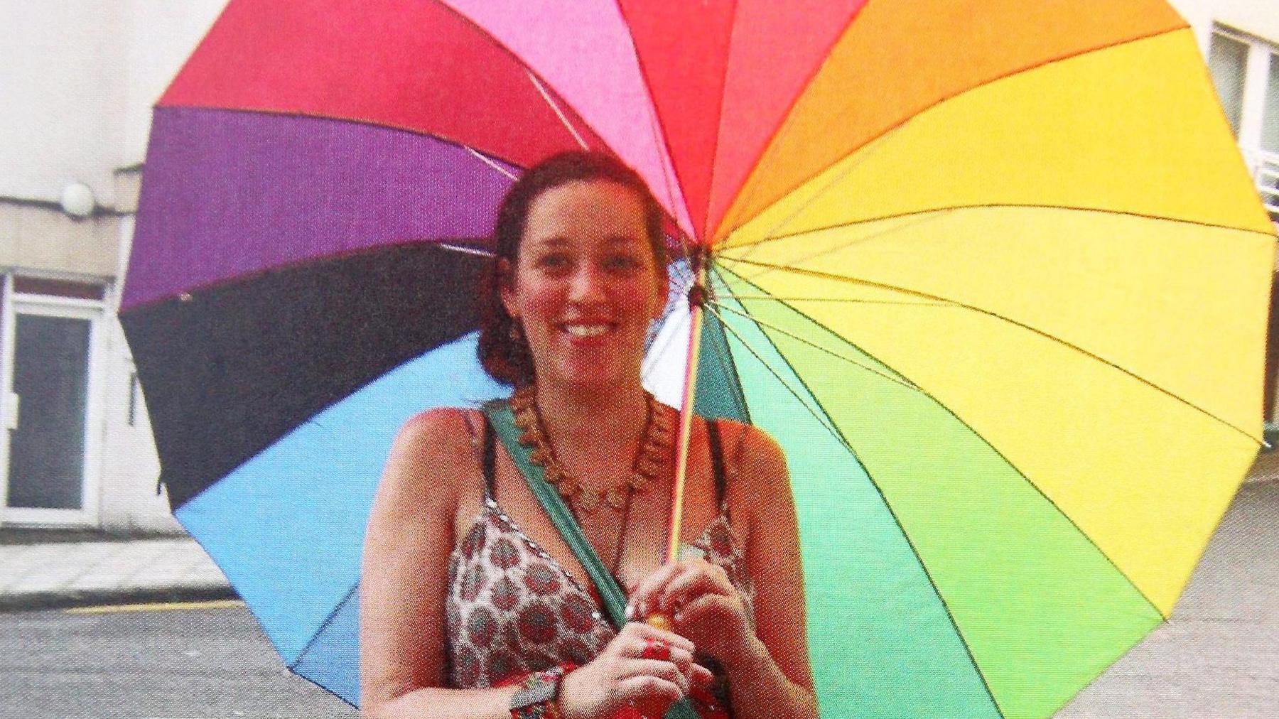 A woman looks at the camera. She is holding a rainbow umbrella and is wearing a cotton strappy top with a wooden necklace and wooden bracelet. The strap of a green shoulder bag is visible.