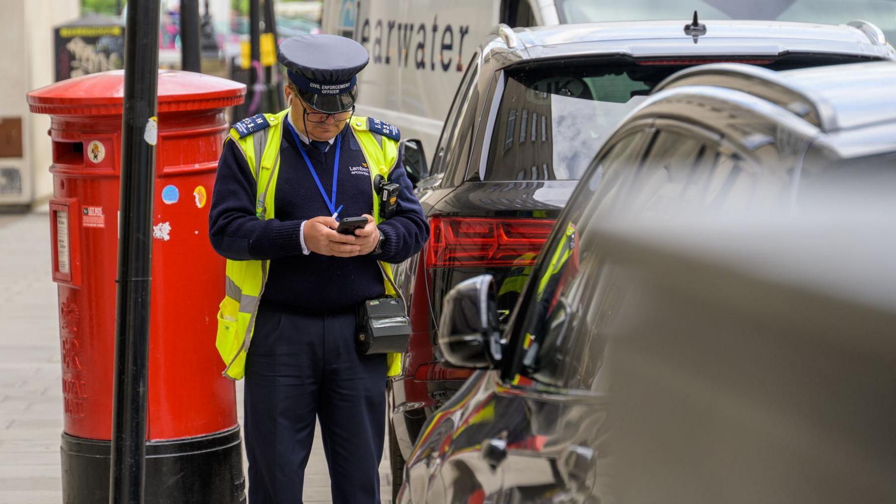 A uniformed traffic warden in a London street is standing by some parked cars and filling out a ticket.