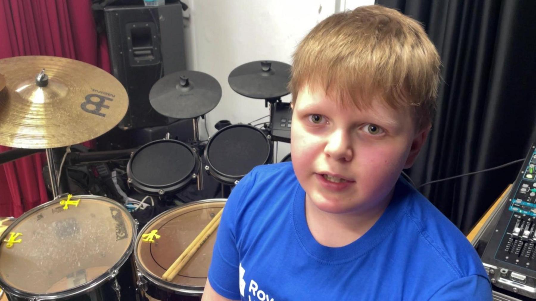 Jack is looking at the camera as he sits at his drum kit. He is wearing a blue T-short and is facing away from his drum kit. A small part of a mixing desk is visible in the bottom right of the image.