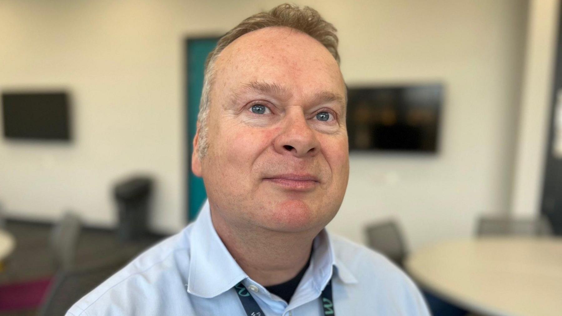 A picture of Dr David Sprake at Wrexham University, he is wearing a pale blue shirt with the top button undone and a black T-shirt underneath. The top of a lanyard can be seen around his neck