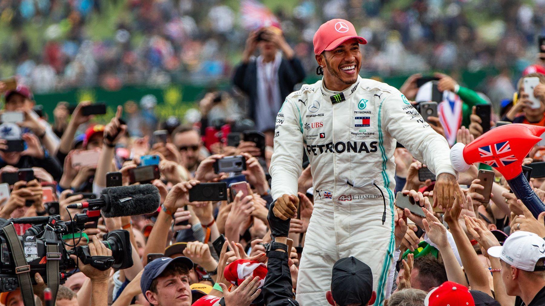 F1 driver Lewis Hamilton wearing a red cap and a white driving suit as he's held up high by a crowd