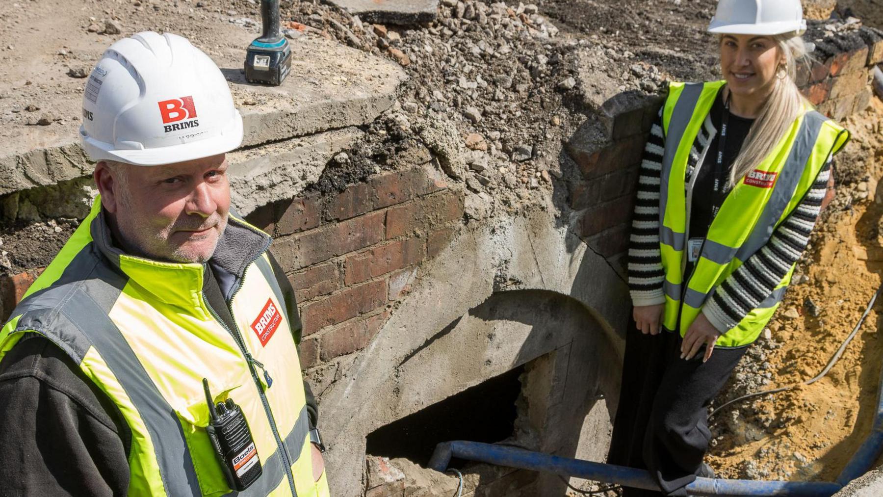 Jason Harrison and Lauren Amerigo are standing either side of a cracked open entrance into an underground air raid shelter. Both are wearing white helmets and yellow high-vis vests.