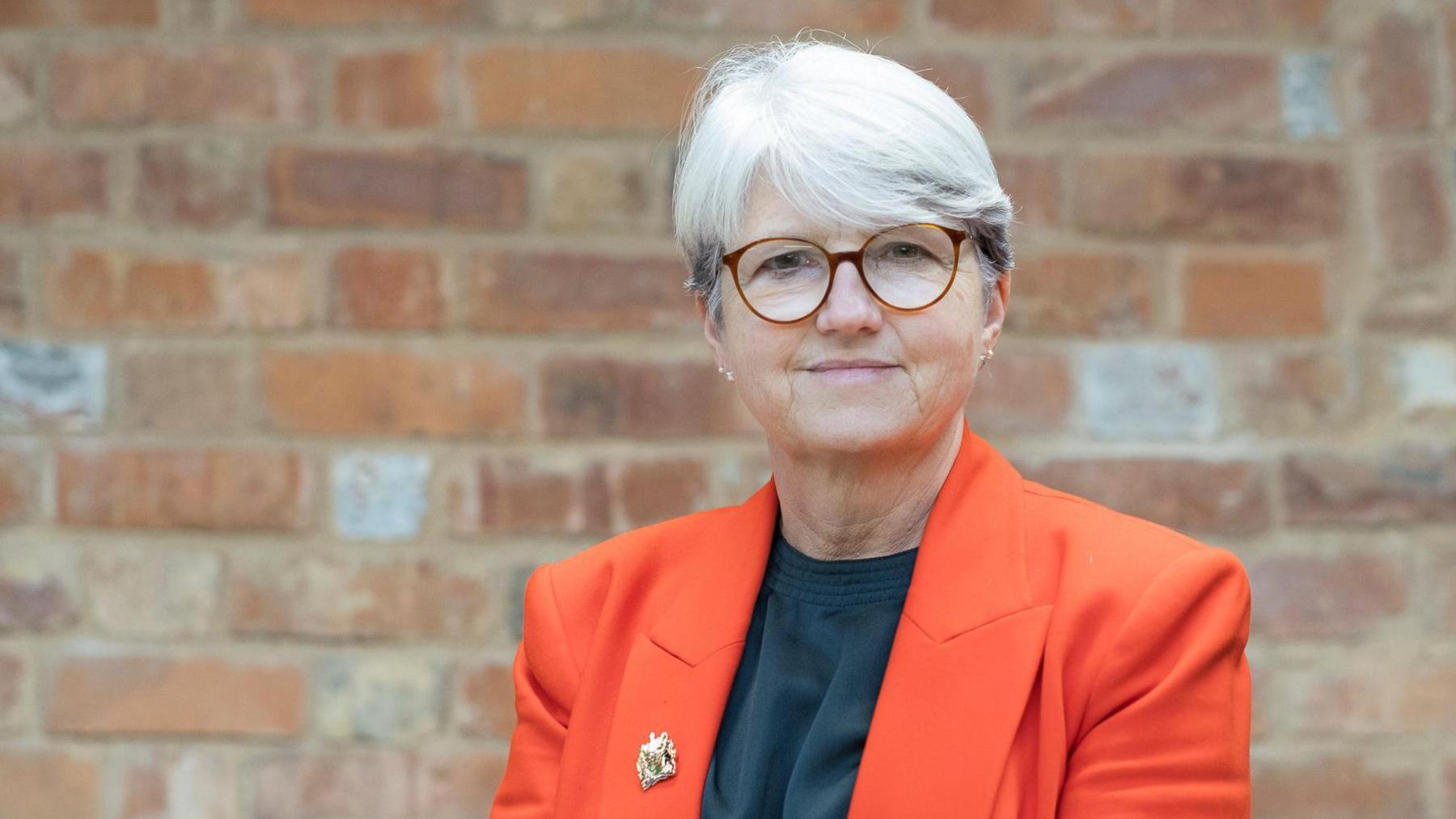 Anna Earnshaw with short white hair and brown-framed glasses wearing a dark top and red jacket with a small broach on the right lapel. She is looking at the camera and standing in front of a brick wall.