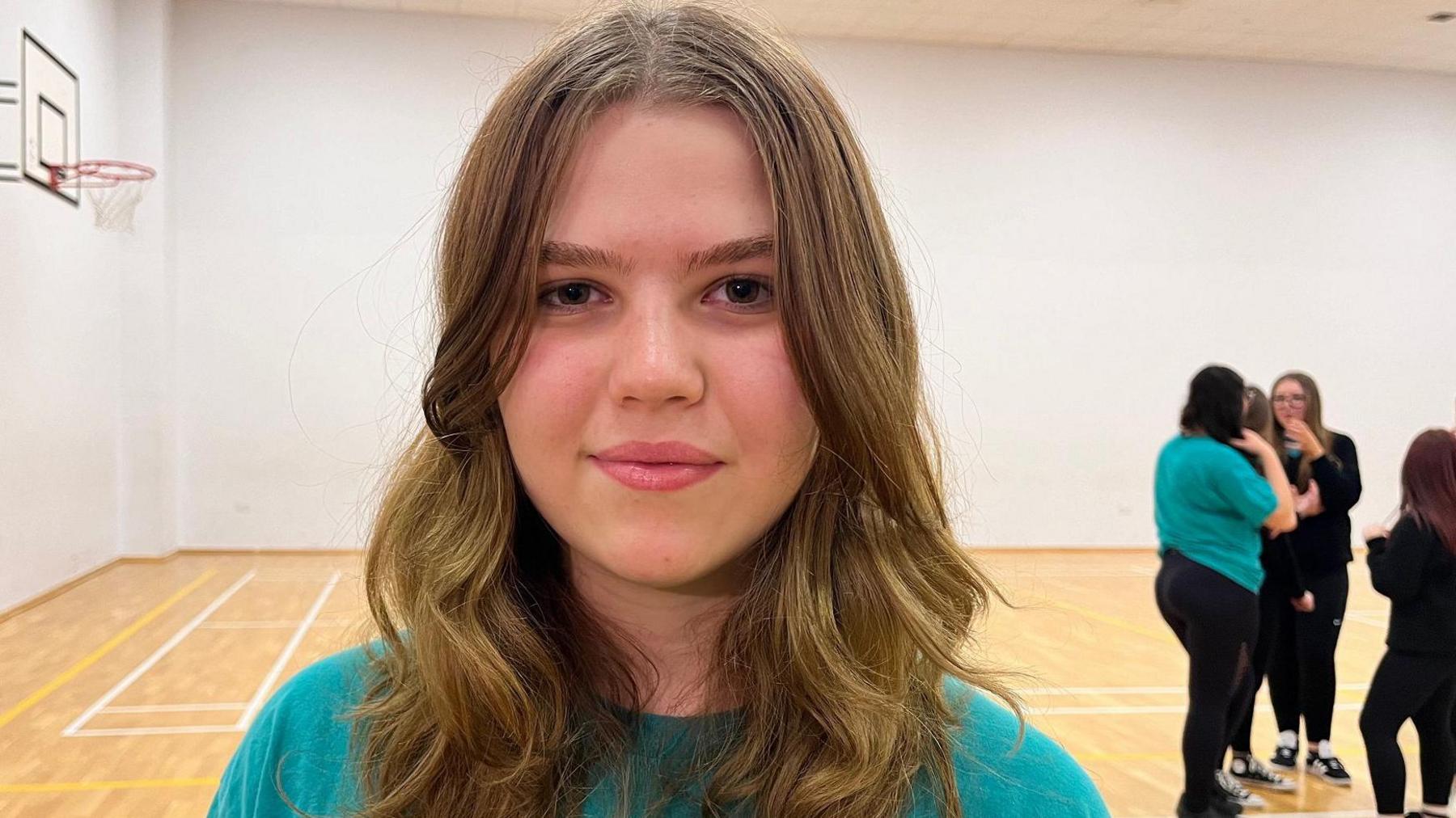 Matilda, a 15-year-old girl, looks into the camera as she stands in a white-walled school gym in front of classmates. She has long, light-brown hair and wears an aquamarine T-shirt.