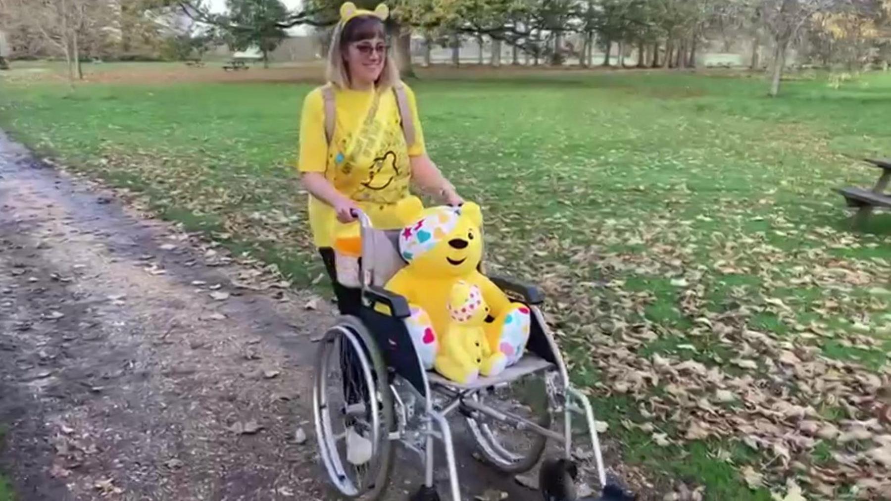 Amy Wright in a yellow CIN t-shirt pushing two Pudsey Teddy bears in a wheelchair in a park