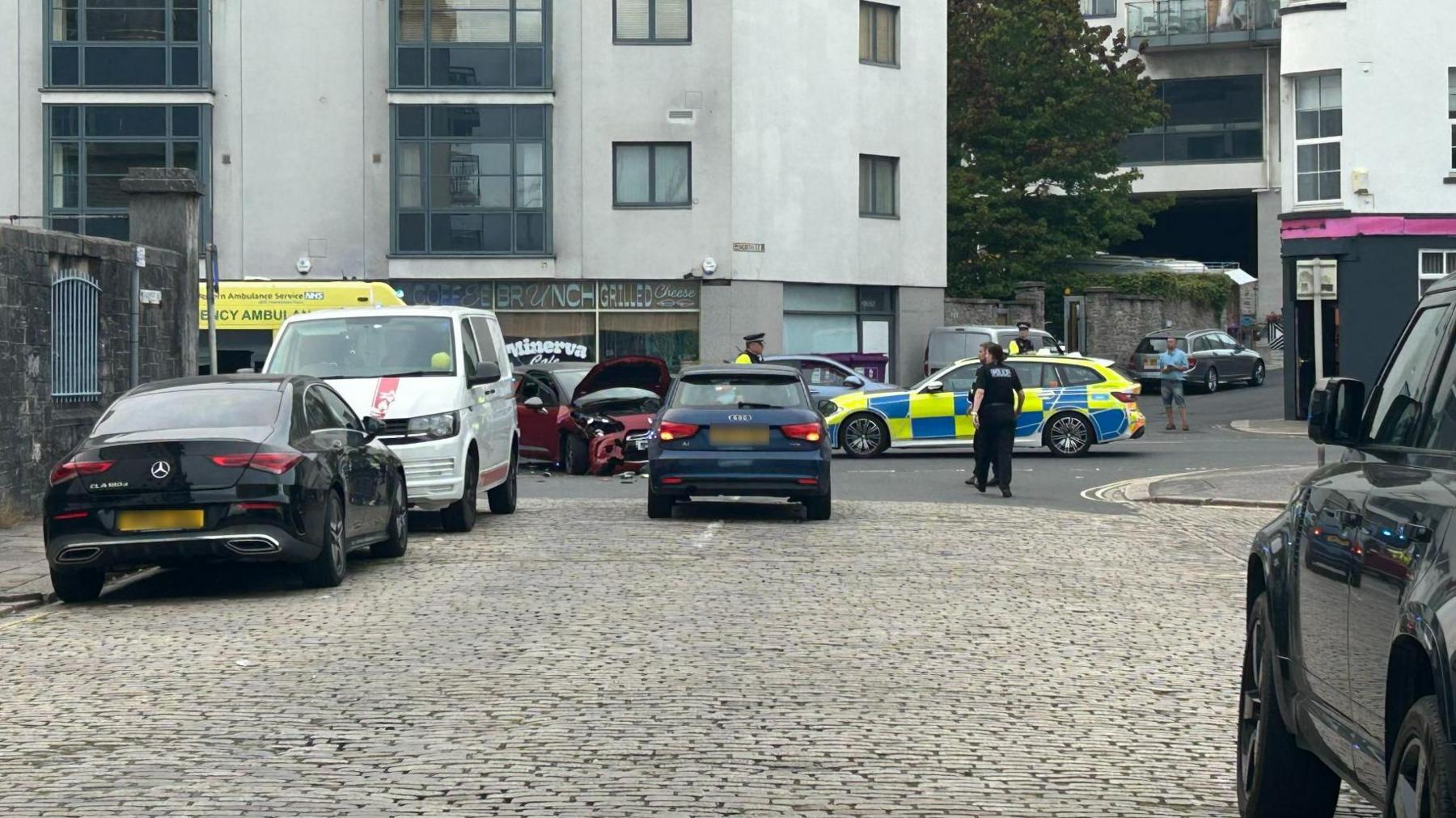 Several Devon and Cornwall Police officers patrol the scene of a crash involving a motorbike and car. The motorbike cannot be seen but the red Citreon DS3 involved in the incident is on the road with significant damage to its front. A police car is parked on the road along with an ambulance.