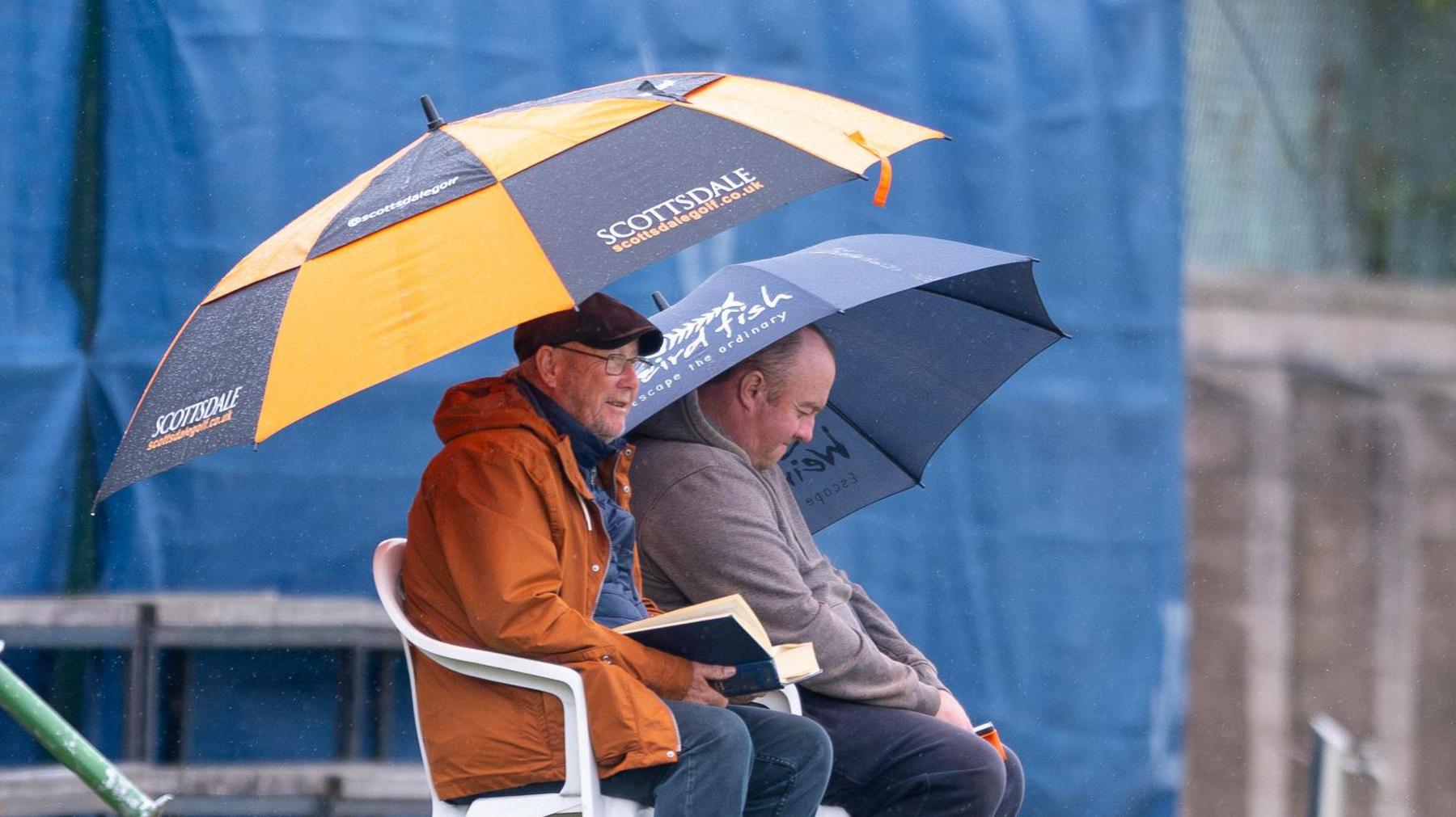 Two spectators hold up umbrellas in the rain at Forfarshire Cricket Club