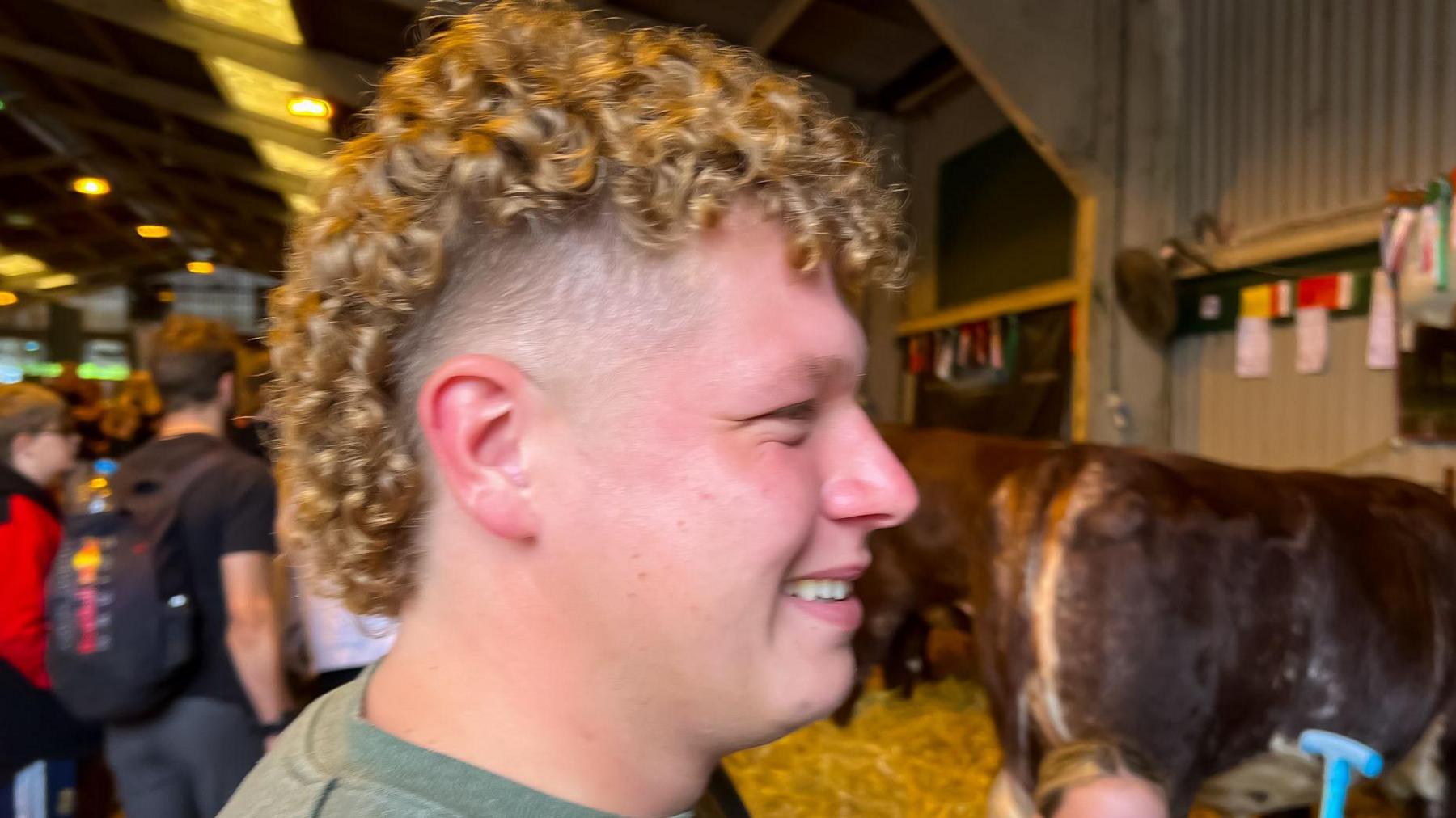 Smiling man in a cow shed with long curly blonde hair on top and in the back and shaved at the sides 