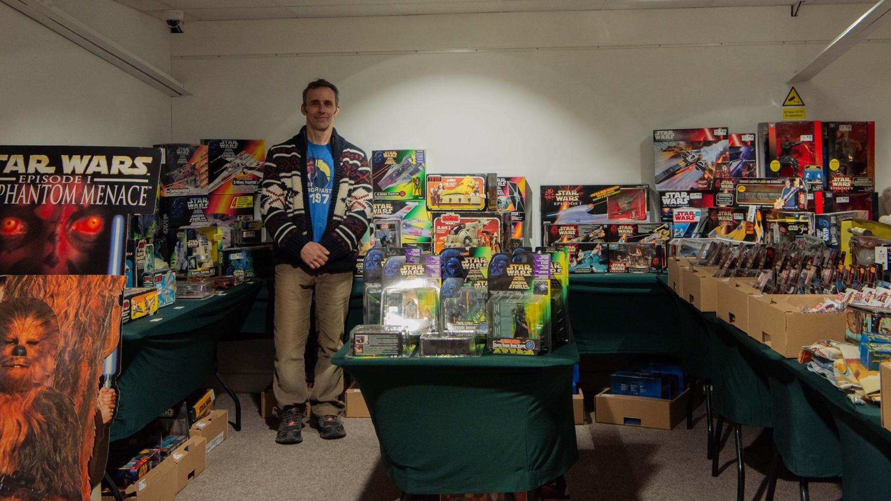 Wide angle shot of Jason Darling, a tall man with dark hair and sideburns, surrounded by tens of boxes of action figures and vehicles from the series at the auctioneers before the collection is sold.