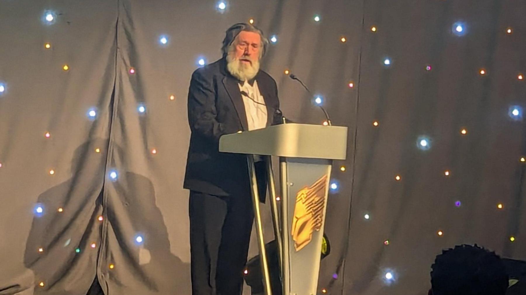 A man in a dinner jacket, white shirt and black bow tie standing behind a black lectern, speaking into a microphone. There are black curtains behind him with multiple spot lights. Tomlinson has a full white beard and long greying black hair.