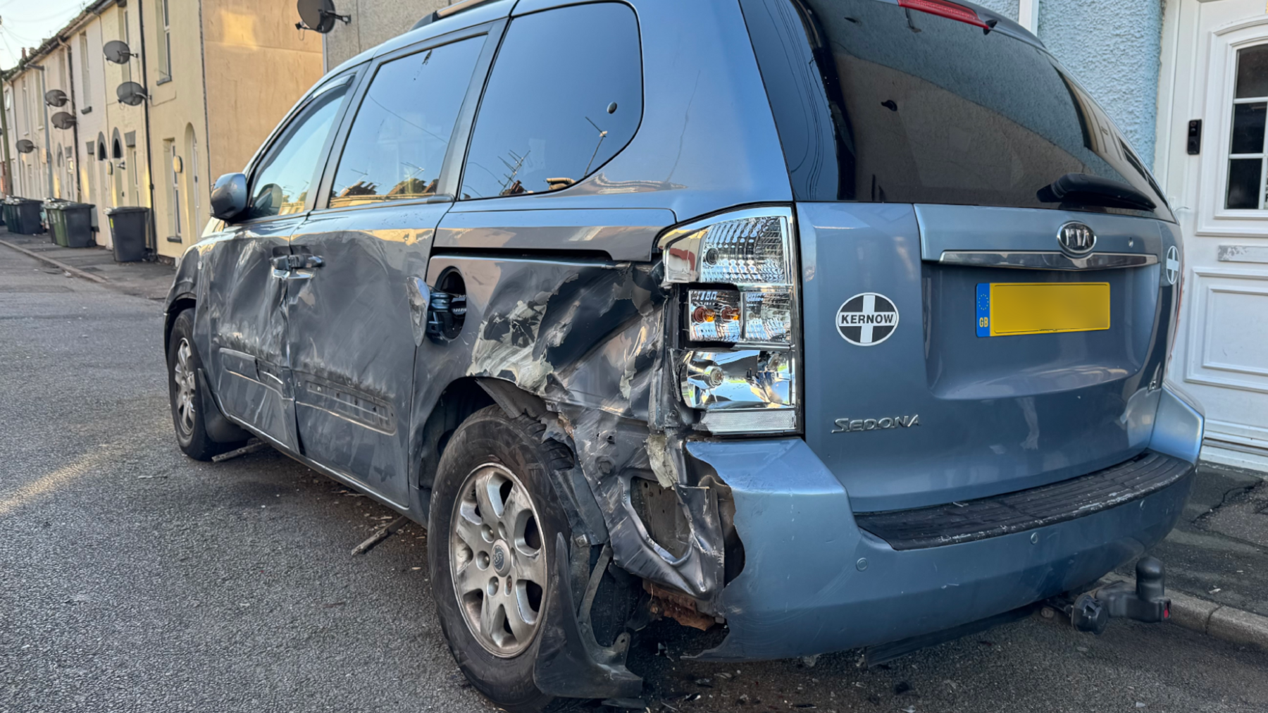 A blue Kia Sedona, with significant damage caused to the near side, caused by a JCB being driven through the street following a raid on a cash machine at a filling station nearby. There are terraced houses on the side of the road, forming the backdrop.