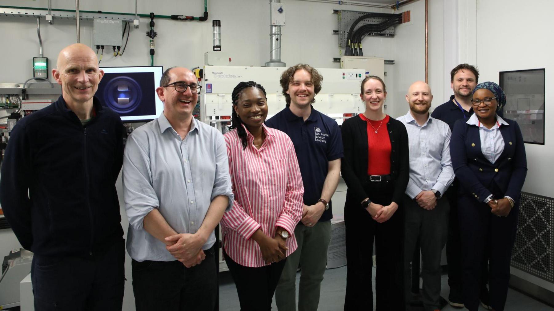 Eight people stand in front of equipment in a lab. There are five men and three women. They are all smiling at the camera.