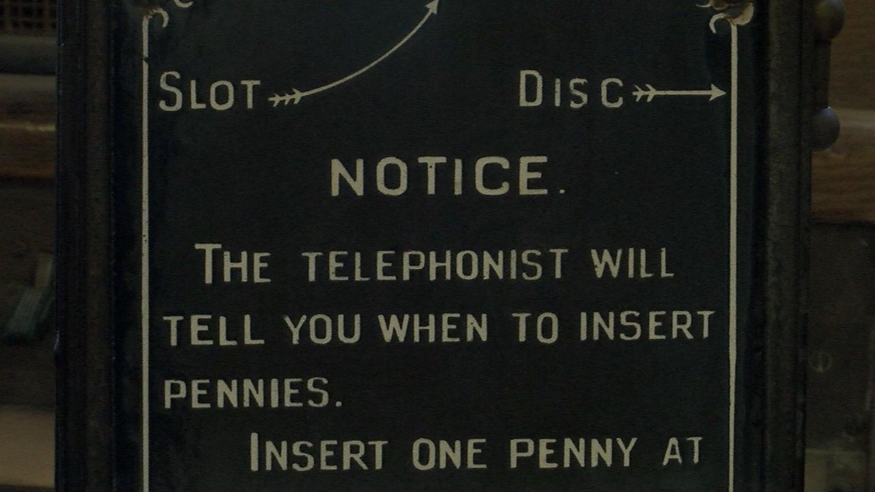 Part of an old payphone. On the black front panel, instructions are written in cream-coloured capital letters. They read: "Notice. The telephonist will tell you when to insert pennies. Insert one penny at [a time]." The place to put money is marked, "Slot".