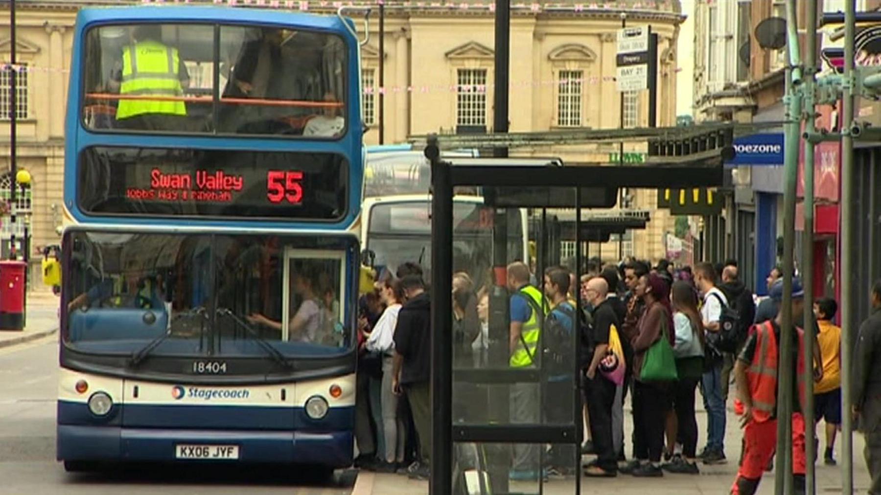 A bus filled with people is parked next to a bus stop. There is a large crowd of people waiting to board the bus.