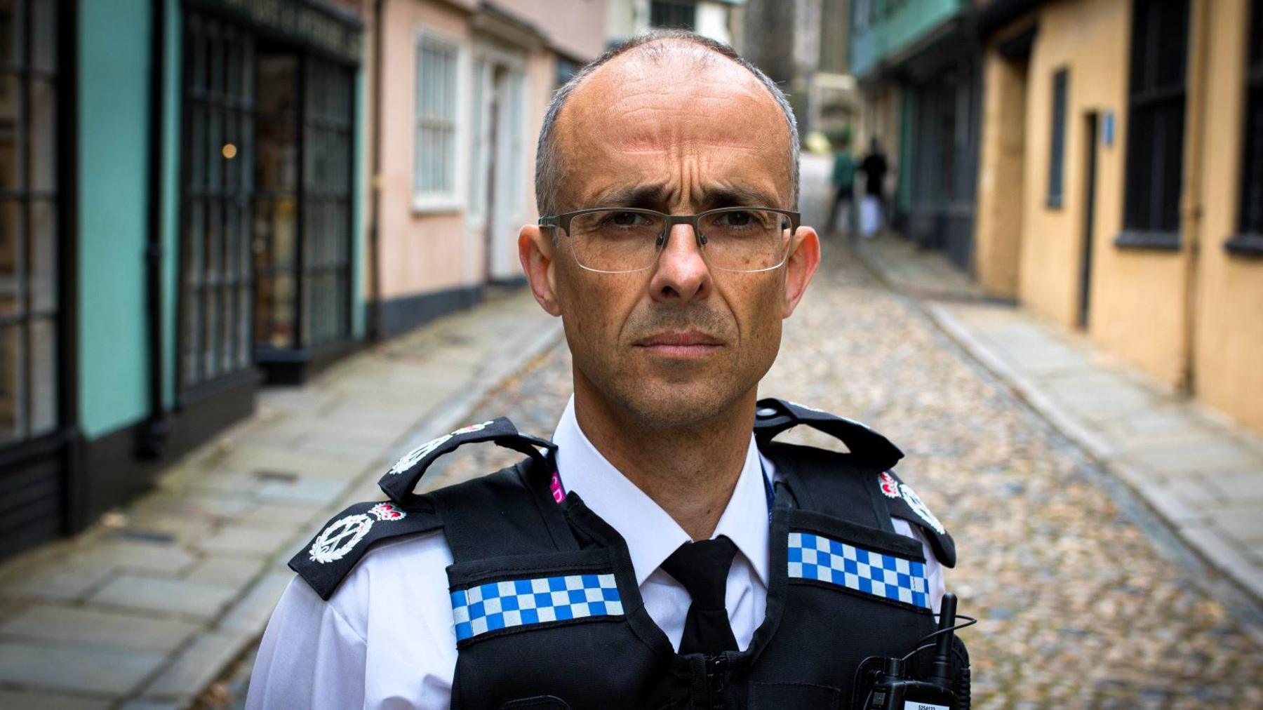 Chief Constable Paul Sanford standing on a small quaint street. He has his uniform on and has short grey hair and glasses. 