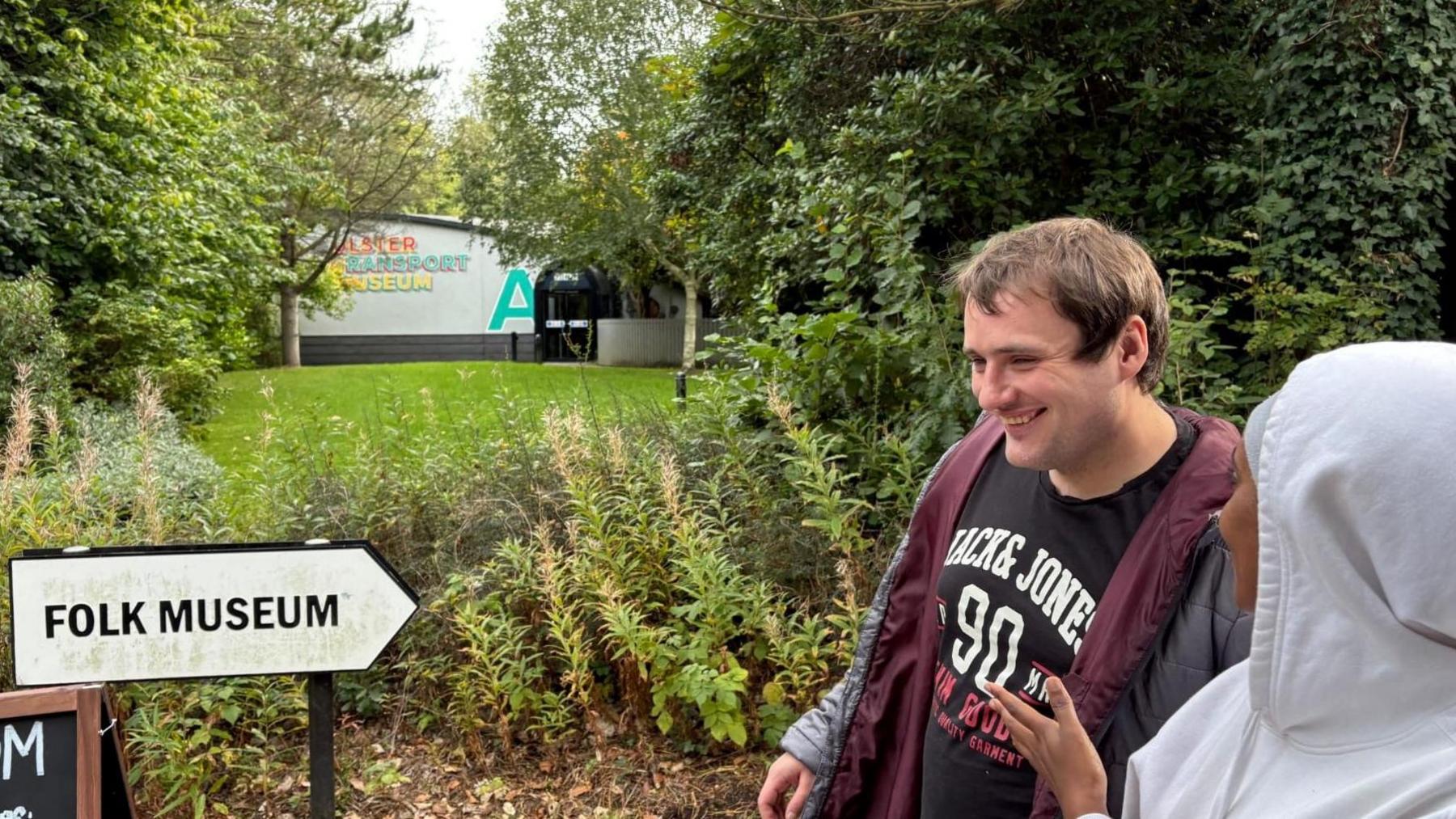 A man and a woman outside at the Ulster Folk Museum. There are trees and grass behind. There is a sign that says 'folk museum' and a visitor centre in the distance. 
