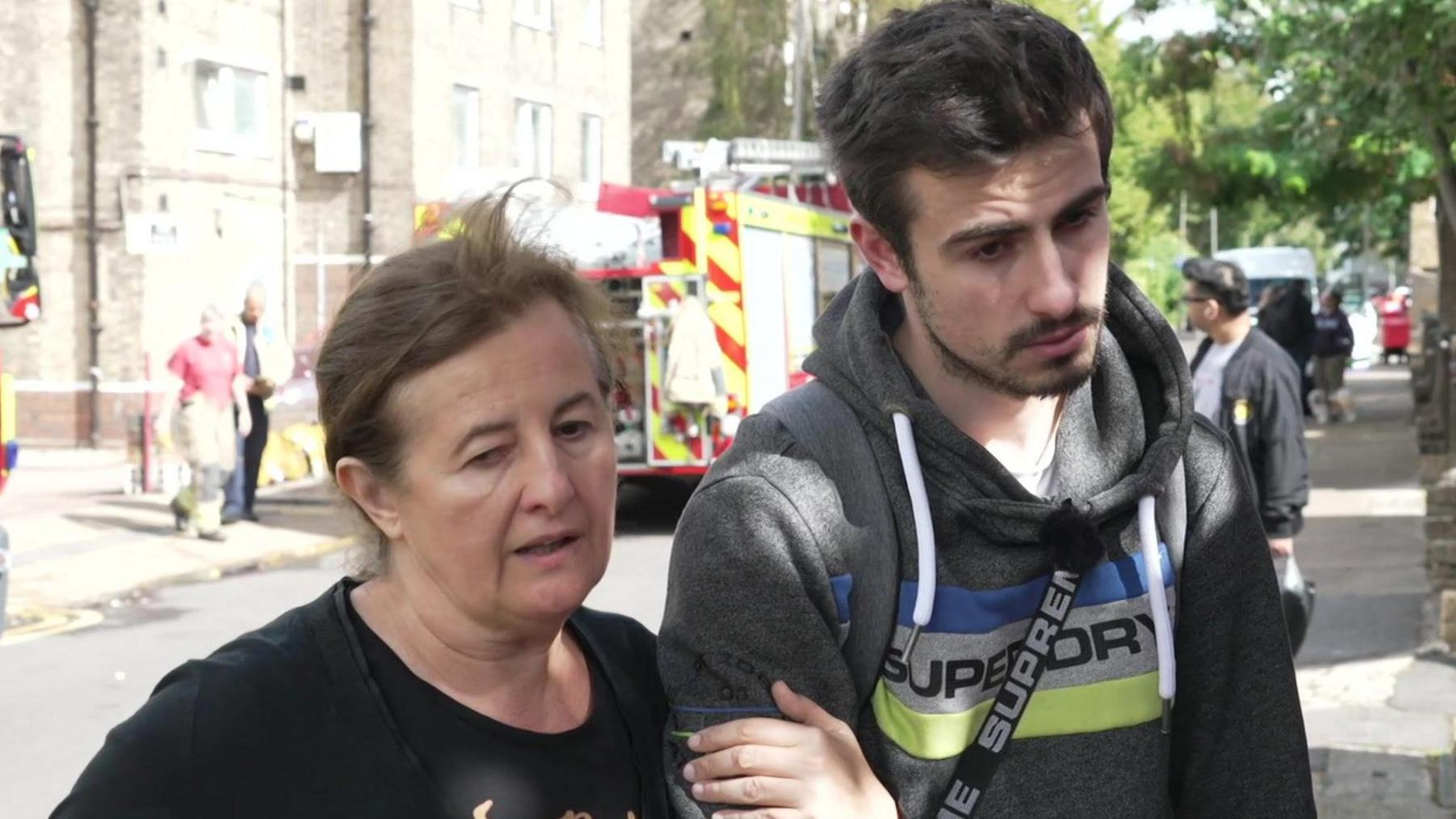 A middle aged woman in a black top stands in the street holding onto her adult son's arm. There are fire trucks in the background. Her son has black hair and a short black beard and is wearing a dark grey hoodie with a blue and yellow logo on the front.