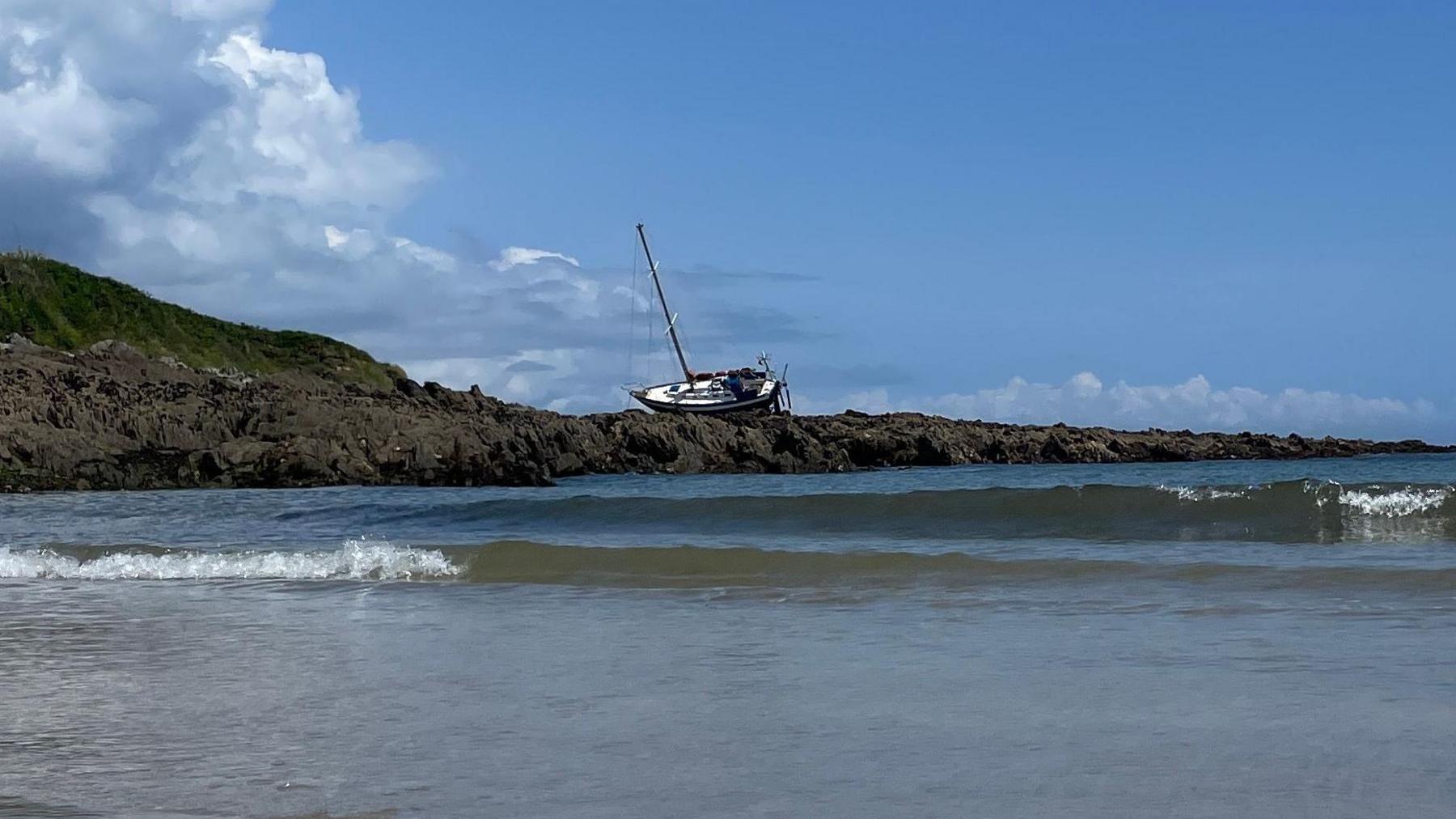 A yacht run aground on rocks along the Cornish coast. Waves are heading to the shore. It is a sunny day with white clouds in the sky.