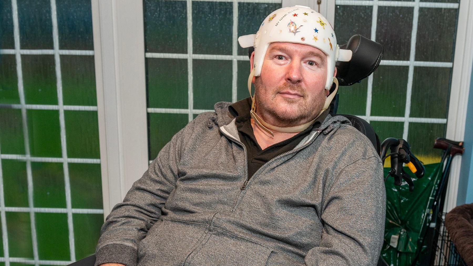 A man wearing a grey tracksuit top sits in a wheelchair in front of closed French doors, with a medical helmet which commonly protects the head when experiencing seizures