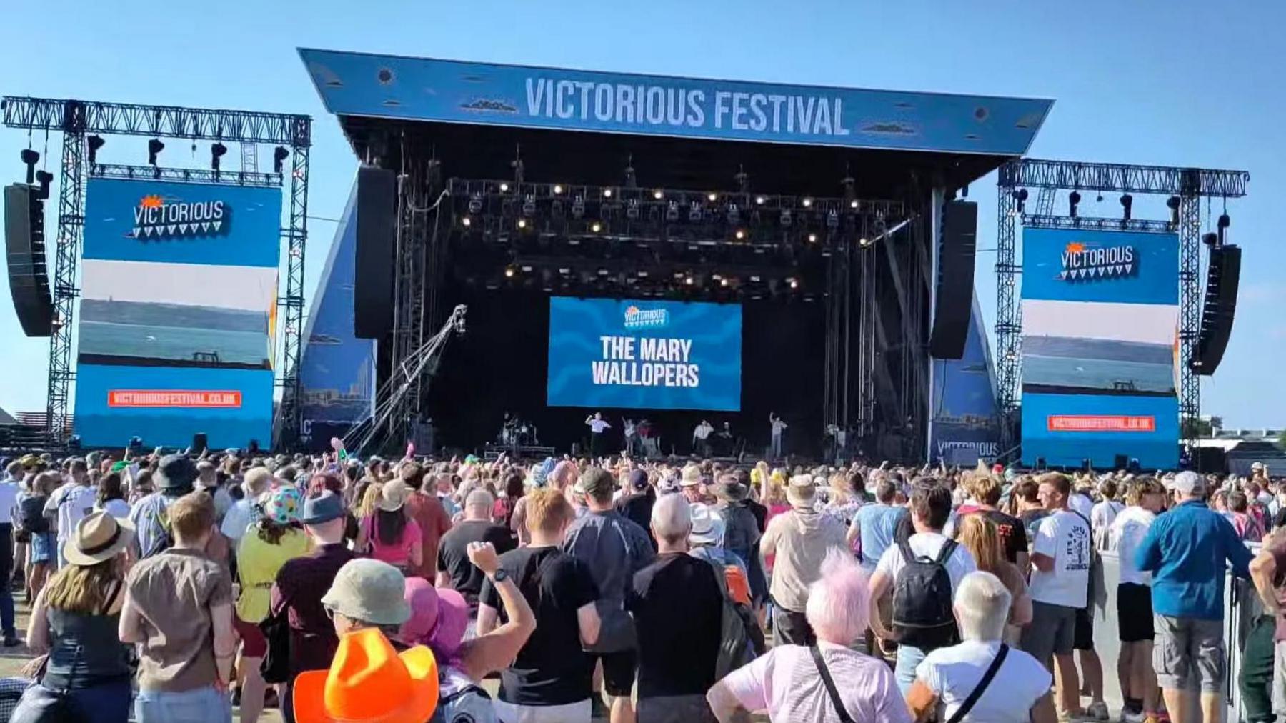 A festival stage with hundreds of people in front of it and The Mary Wallops written in large letters behind the band