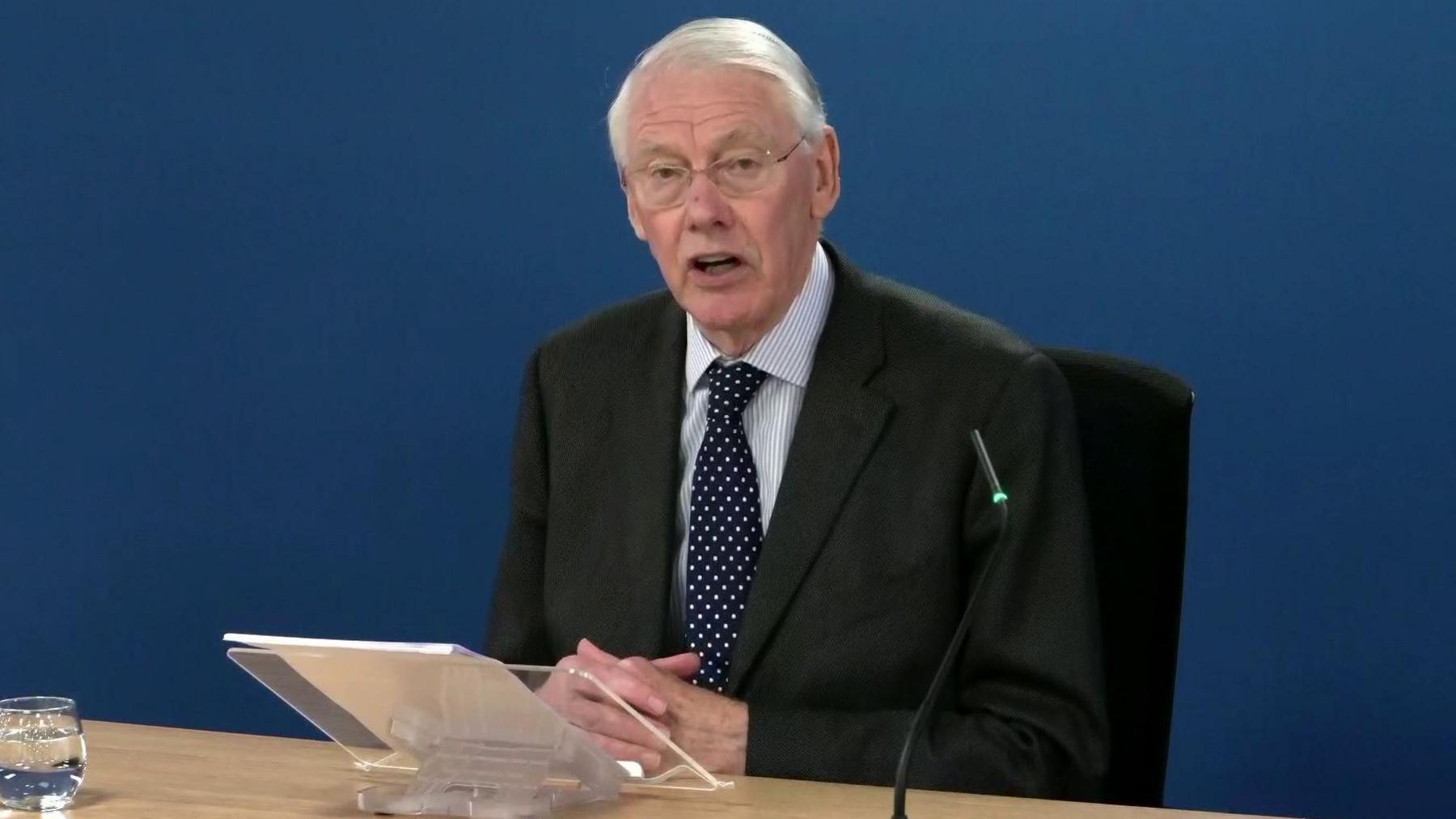 Image shows Sir Martin Moore-Bick, a Caucasian man with white hair and glasses, wearing a black suite with striped shirt and a blue tie with white spots, sitting at a desk against a blue backdrop