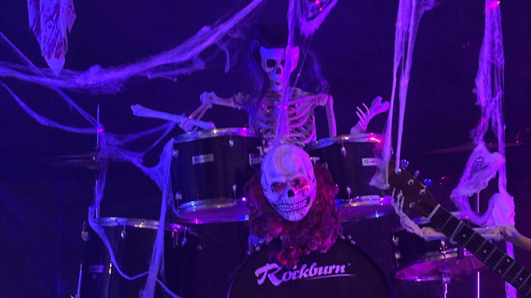 A skeleton playing on a drum kit with a skull attached to it. There is the top of a guitar on the right hand side of the image and cobwebs surrounding the scene which is set against a black back drop.