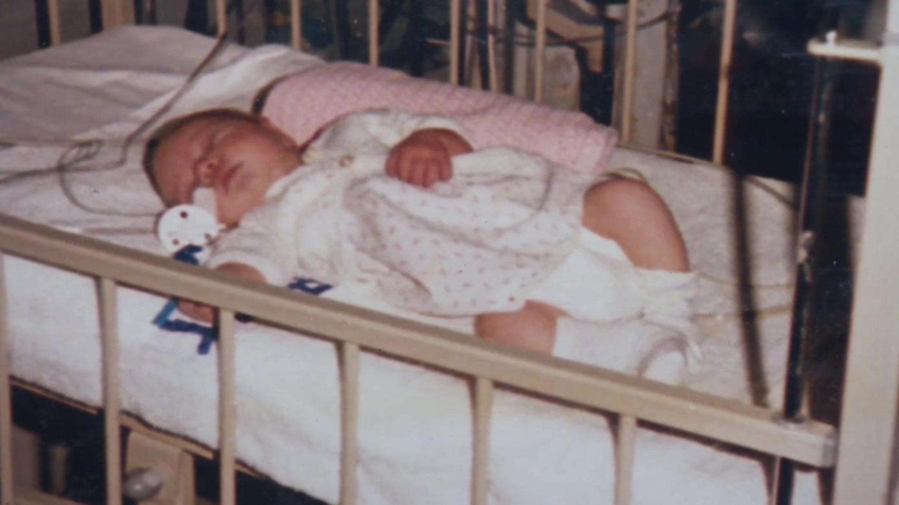 A slightly blurry picture of Natalie Walsh as a baby wearing a white dress and lying in a cot 