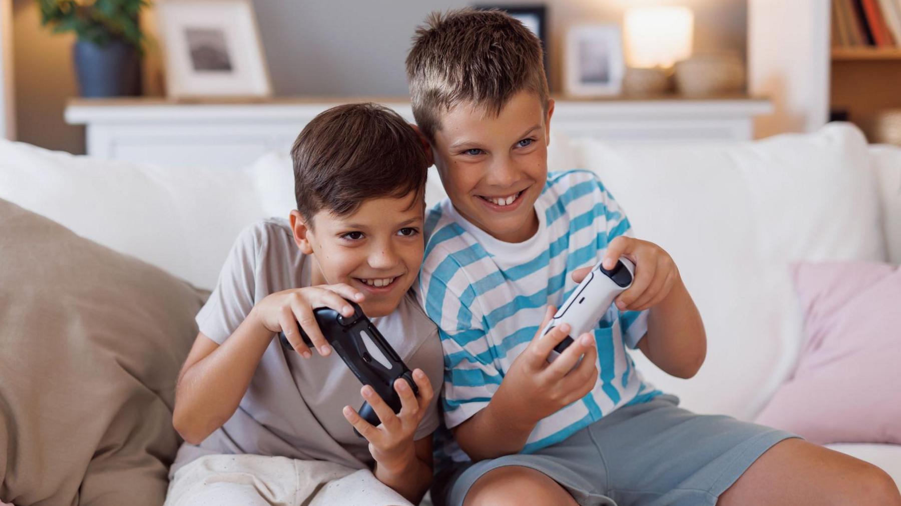 Two boys are sitting close together on a white sofa, holding game controllers and smiling while playing video games in a cozy living room filled with plants and decor.