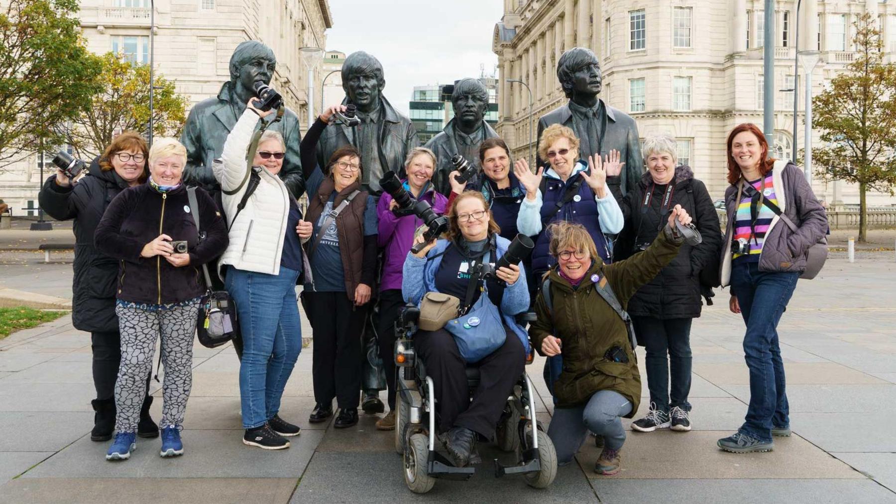 A group of women photographers pose for a picture in Liverpool, in front of a statue of The Beatles. They are holding their own cameras in the air. One of them is a wheelchair user.