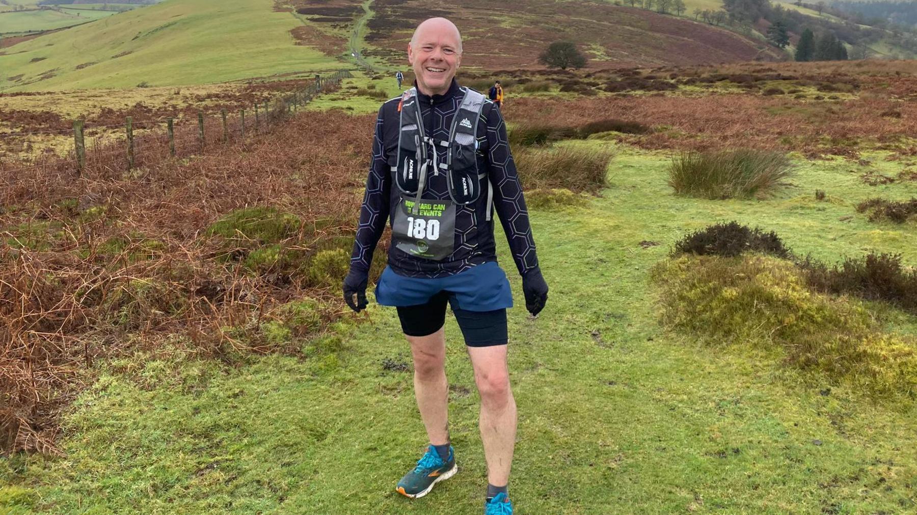 A man with a shaved head stands on an expanse of grass and heathland. He is wearing trainers, black running shorts, a black running top and black gloves and has a rucksack on his back. He has a sign pinned to his front with the number 180 on it.