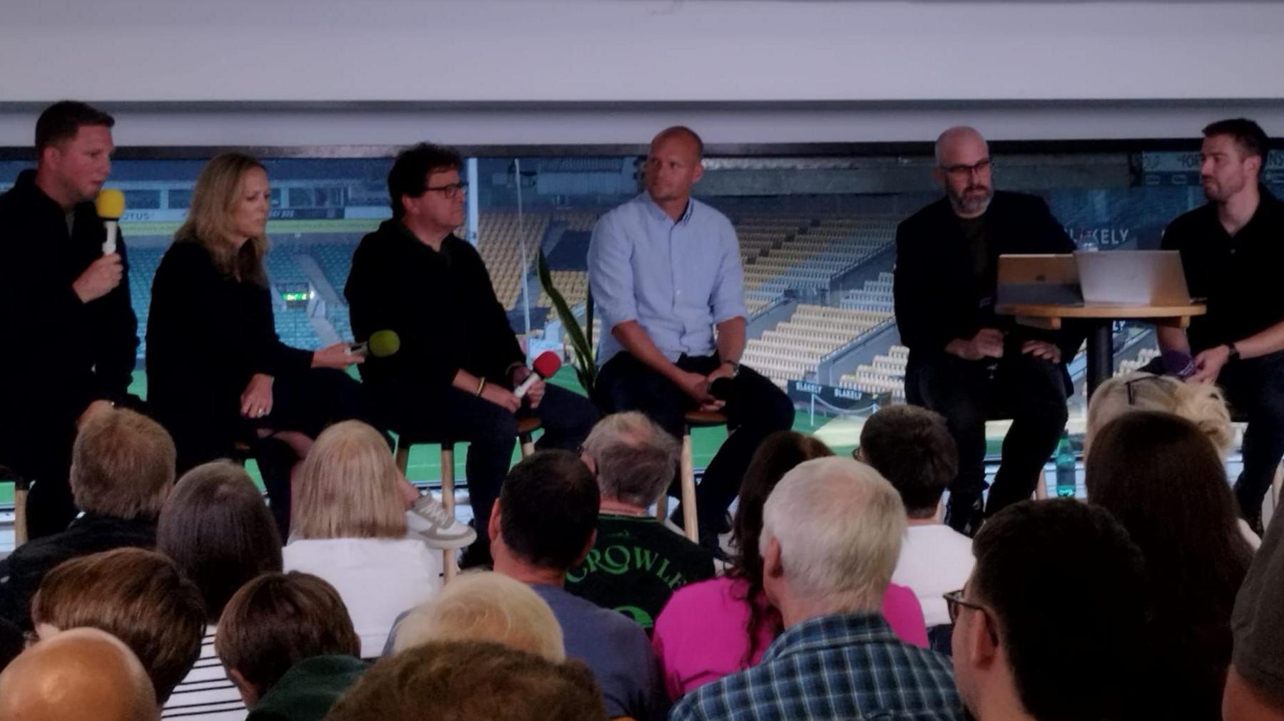 Six people sit on stools with microphones as they speak to a crowd
