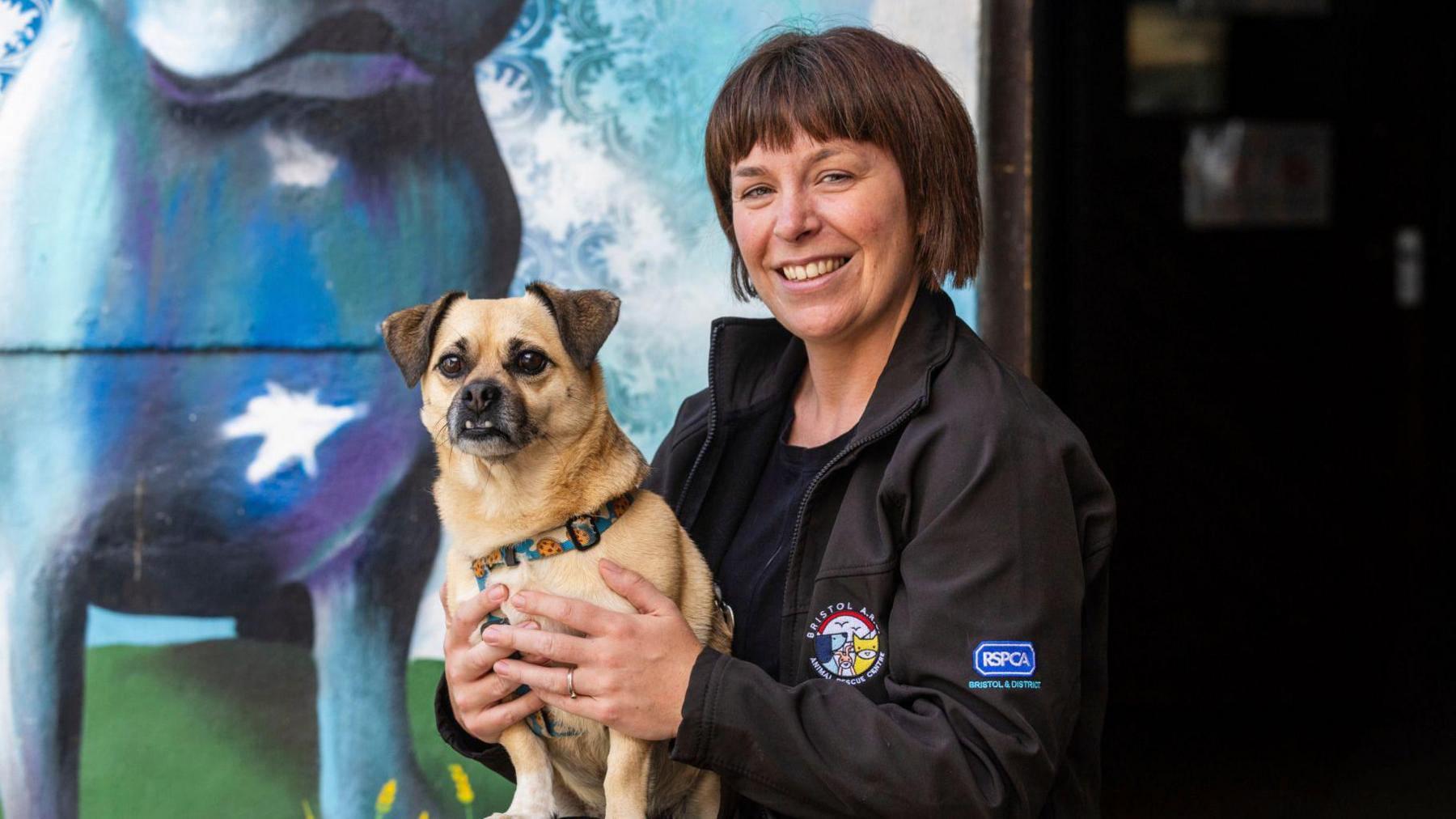 Rosie El-Tibi wearing a black jacket with the animal centre and RSPCA logos on it. She is kneeling down beside a large blue mural of a dog, holding a dog to her chest and smiling. 