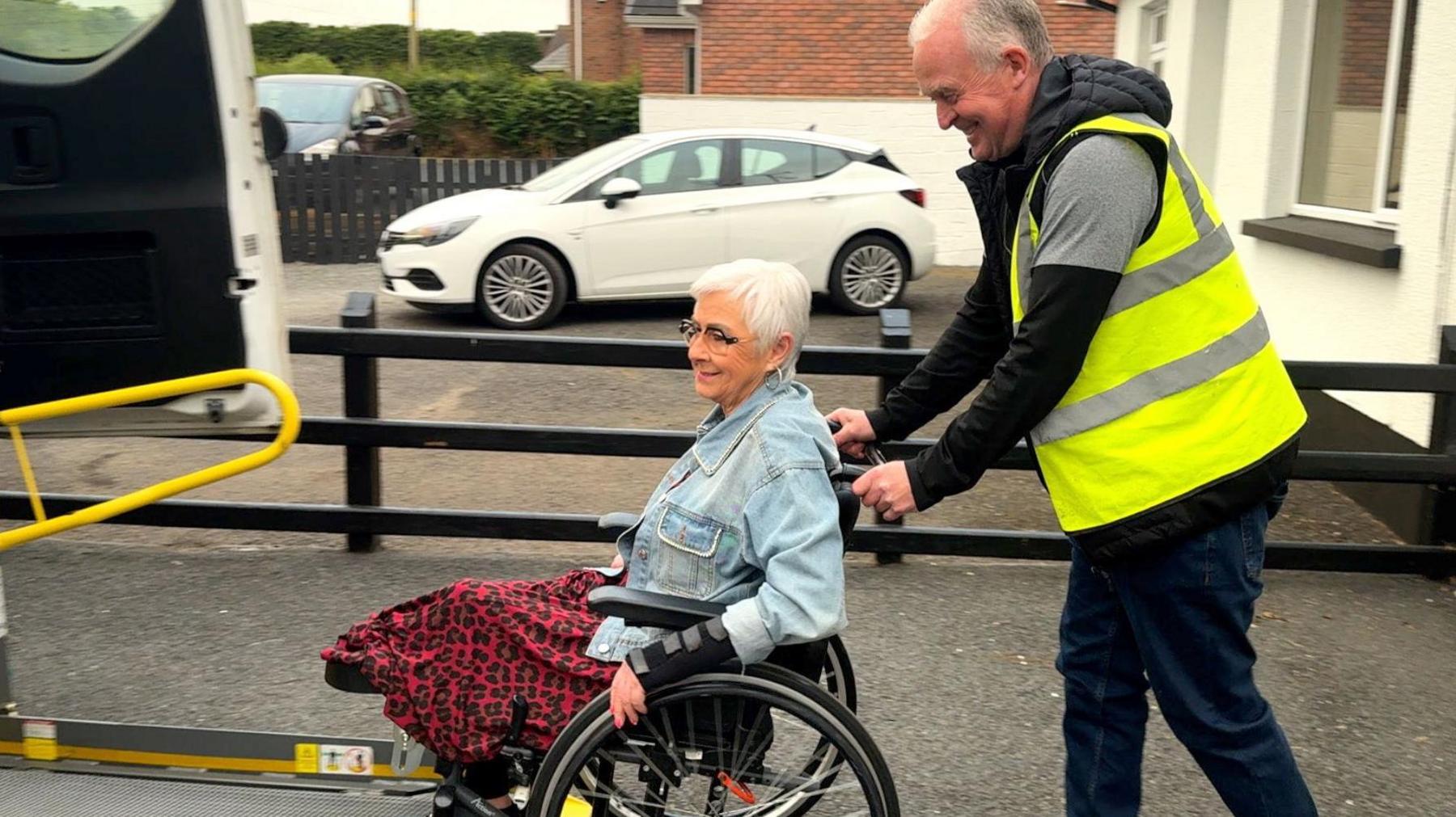 A woman in a wheelchair being pushed up a ramp into an accessible vehicle by a man in a hi-vis jacket. She has short grey hair and is wearing a long patterned red skirt and a denim jacket