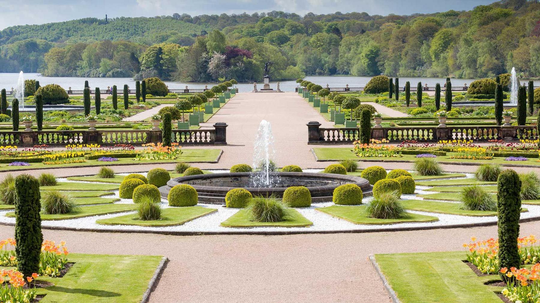 The gardens at Trentham Estate -  an oval with bushes and grass features a water fountain in the centre with a large walkway circling it and leading to a lake in the distance. A forest is visible in the background on the opposite side of the lake.