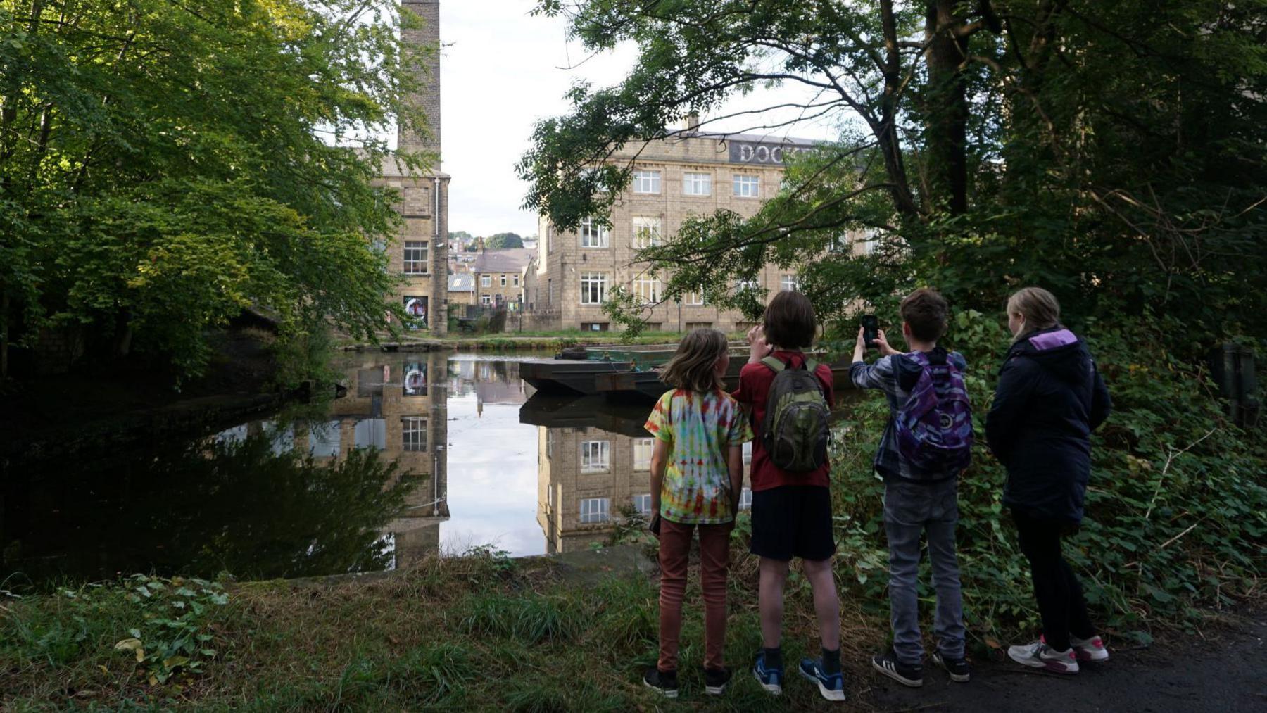 Four young people dressed in casual wear stood on a grass riverside overlooking water. One child is pointing a mobile phone and taking picture. 